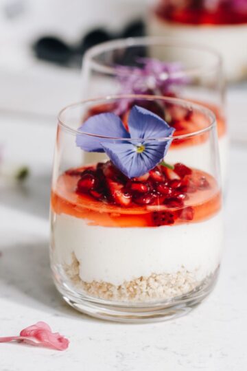 A decorated white chocolate mousse in a glass sits on a white table. In the glass is a biscuit base, a white chocolate mousse and finely chopped berries with juice on it and an edible purple flower on top. Half of another set mousse is behind it. In the far background is a white tray with more desserts in glasses.