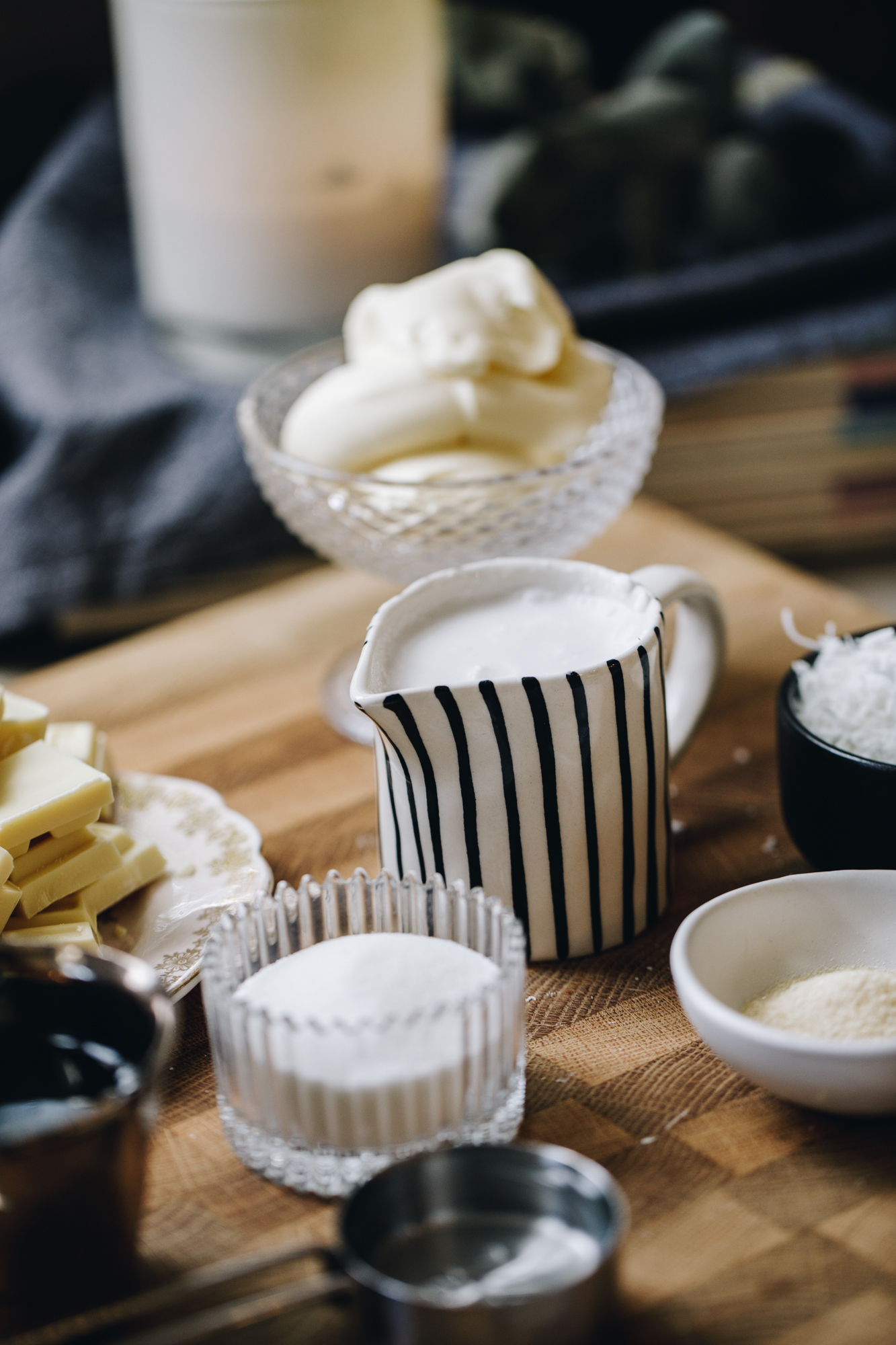 A wooden board sits on a kitchen bench. ON the board is vintage bowls and ramekins with ingredients in it. There is cream, white chocolate, sugar, gelatine powder, coconut and mascarpone in view. In the background is a dark grey tea towel with a large white candle on it.
