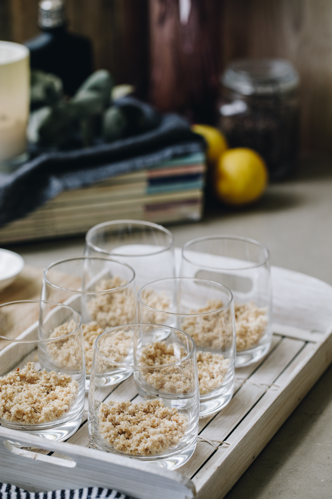 A white wooden tray sits on a black and white tea towel on a brown kitchen bench. On top of the tray is 6 glass that has a biscuit crumb in the base of each one. Behind the tray is a stack of magazine with a blue cloth on them. On the cloth is a white candle and a small black bottle. Lemons are beside the candle and a vintage glass jar.