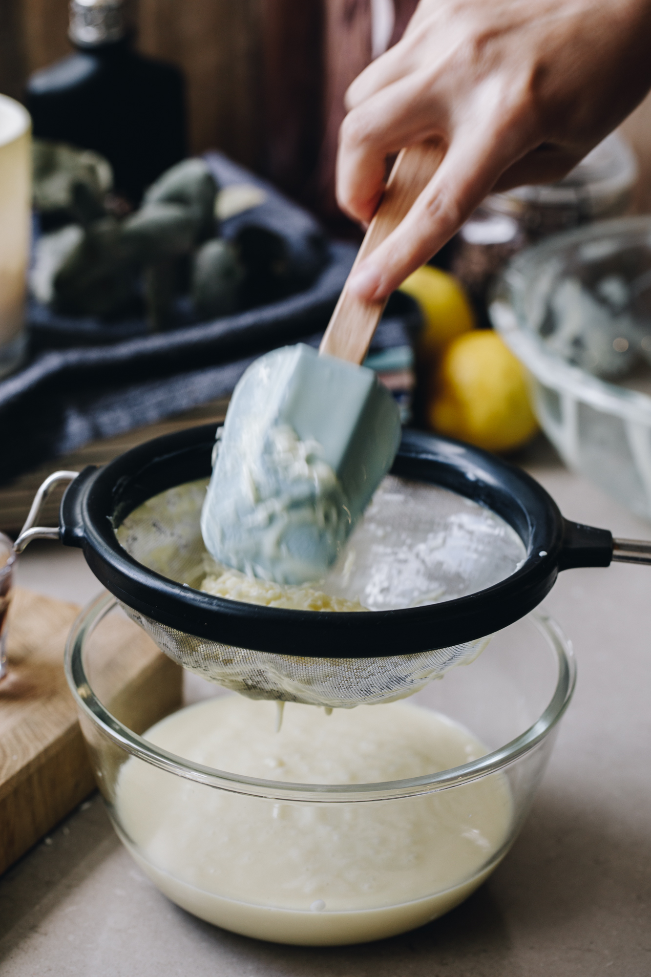 A glass bowl sits on a brown kitchen bench. A white chocolate cream is in the bowl and a black sieve is on top with coconut remnants in it, it is pressed by a blue spatula with a wooden handle. Behind the tray is a stack of magazine with a blue cloth on them. On the cloth is a white candle and a small black bottle. Lemons are beside the candle and a vintage glass jar.