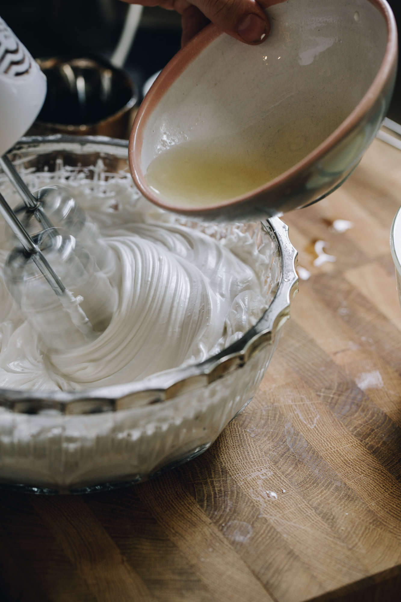 A glass vintage bowl with a silver trim sits on a wooden board. In the bowl is a meringue that is being whipped by a white hand mixer. At the same time Naomi is pouring in the melted gelatine mixture.