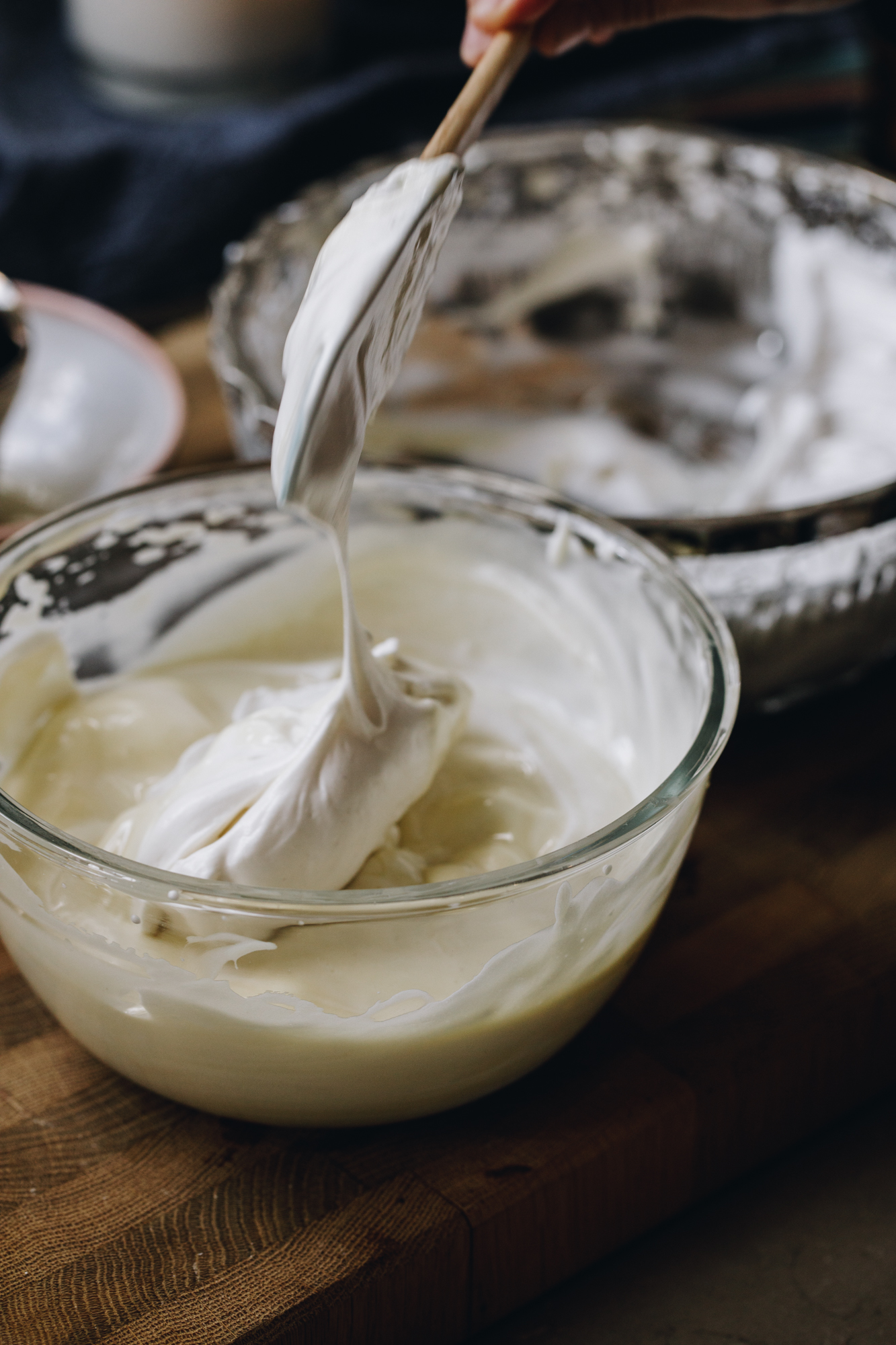 A glass bowl sits on a wooden board with white chocolate cream in it. A spatula is adding a blob of meringue in the centre. Behind it is a glass vintage bowl with a silver trim has remnants of meringue in it.