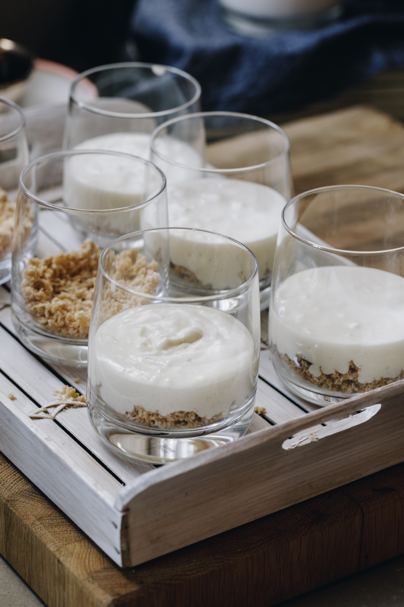 A white wooden tray sits on a wooden board. On the tray is 6 glasses that have sbicuits crumb in all of them and four out of the six have white chocolate mousse on top.