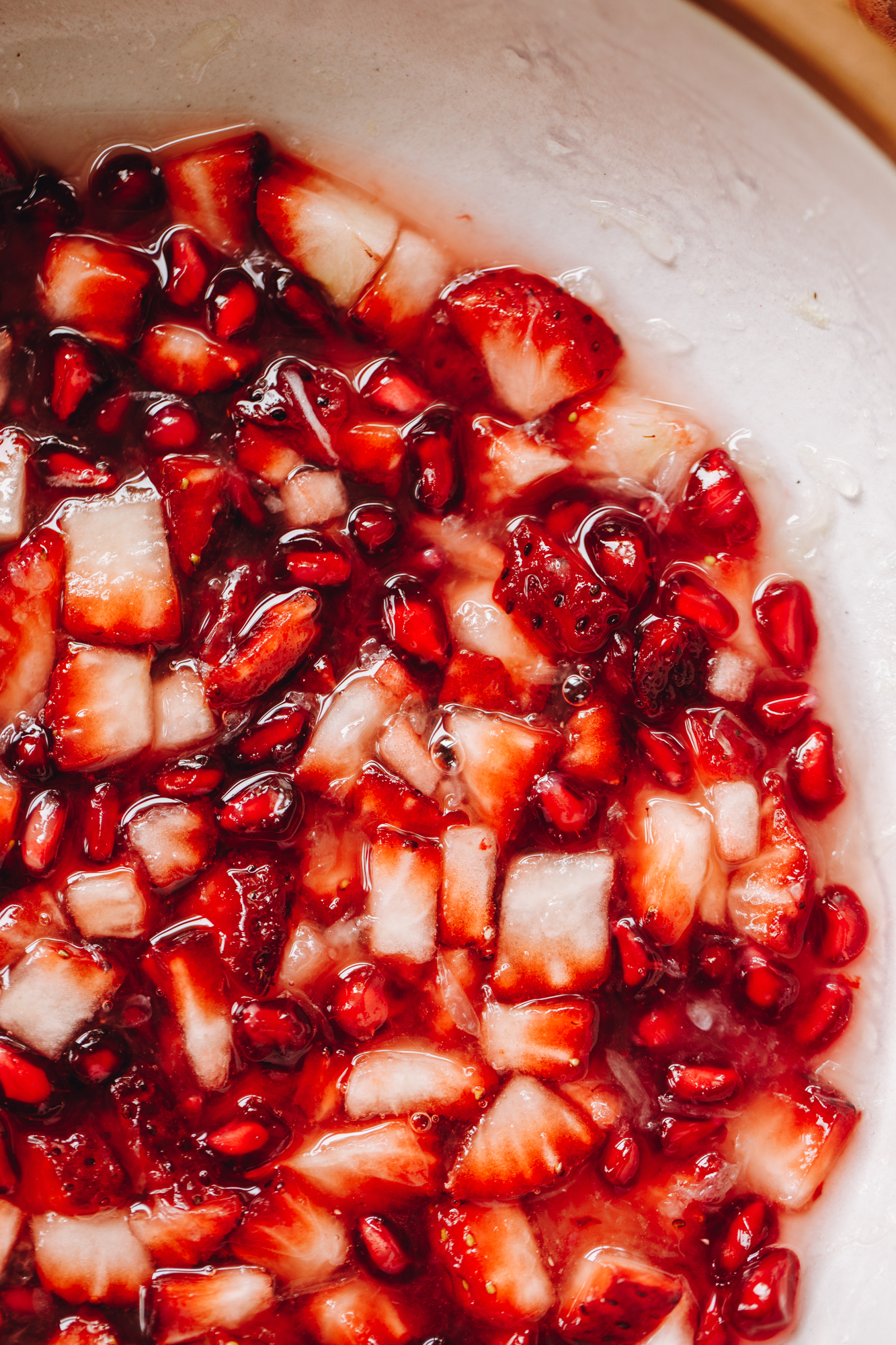 A flat lay shot shows cut up strawberries and pomegranate seeds in a ceramic bowl in a clear juice.