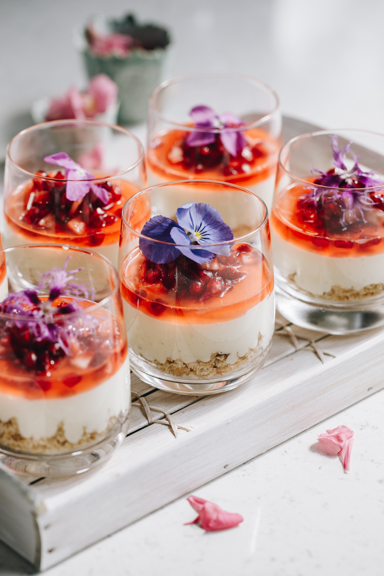 On a white wooden tray sits 6 decorated white chocolate mousse desserts. In the glasses is a biscuit base, a white chocolate mousse and finely chopped berries with juice on it and an edible purple flower on top.