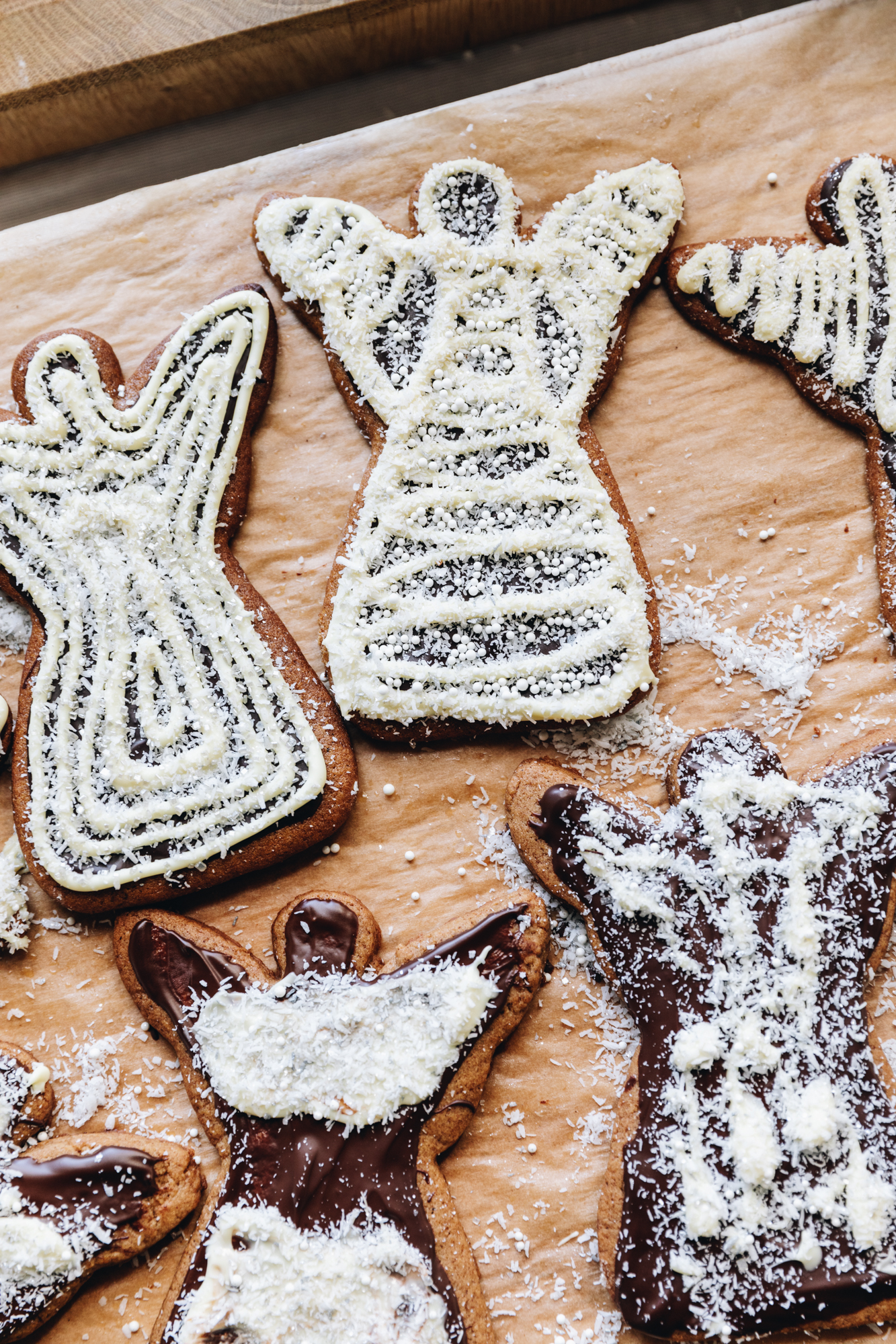 A flat lay shot shows a wooden board line with brown baking paper. On the paper is freshly decorated spiced gingerbread cookies that are shaped as angels and are decorated with dark chocolate and white chocolate and sprinkled with glitter coconut. 