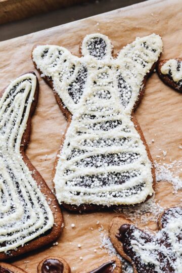 A flat lay shot shows a wooden board line with brown baking paper. On the paper is freshly decorated spiced gingerbread cookies that are shaped as angels and are decorated with dark chocolate and white chocolate and sprinkled with glitter coconut.