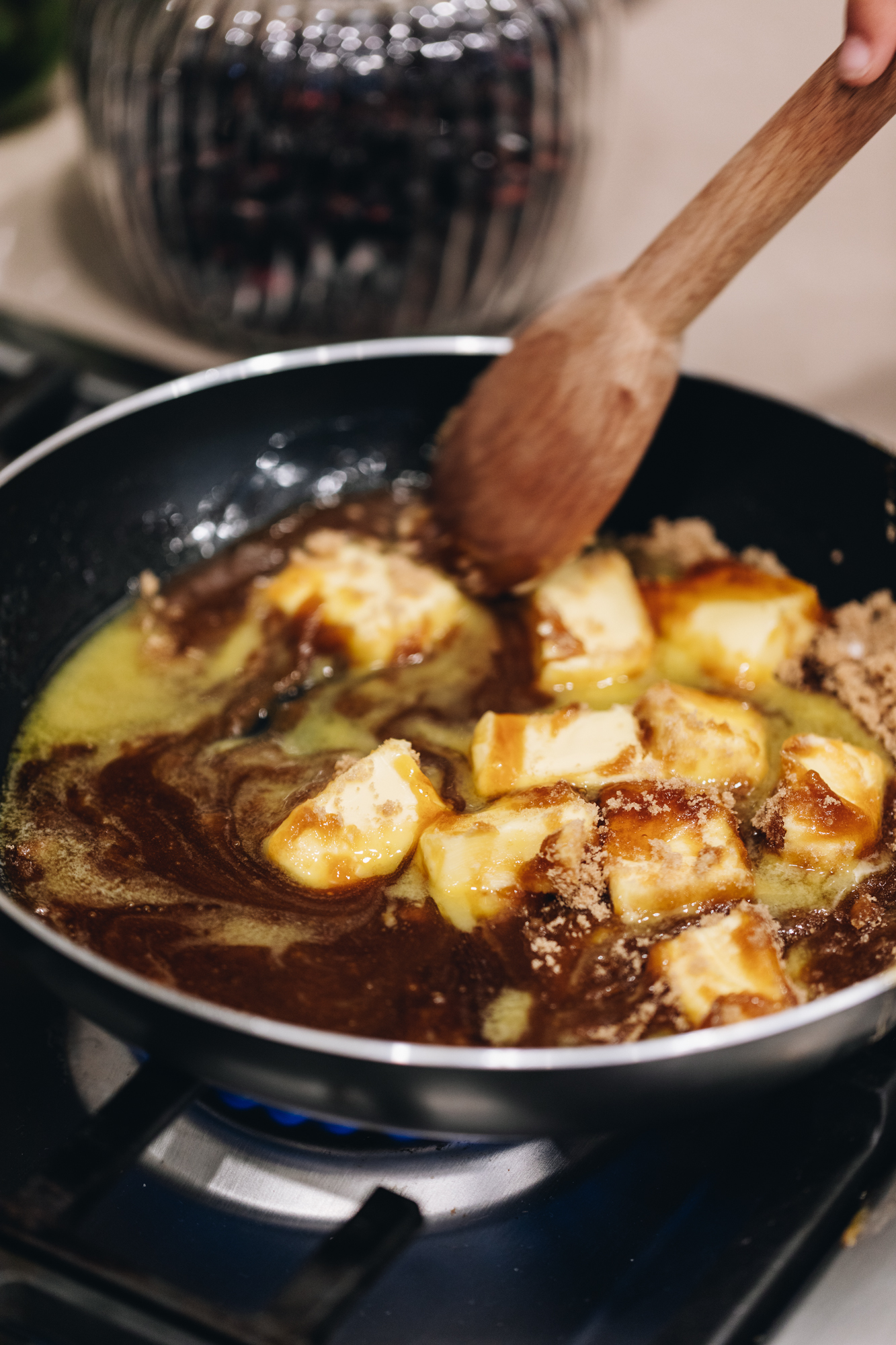A black frying pan is on a black gas hob. In theban is butter, brown sugar and golden syrup that is being stirred by a wooden spoon. A glass jar with a lid is in the background. 