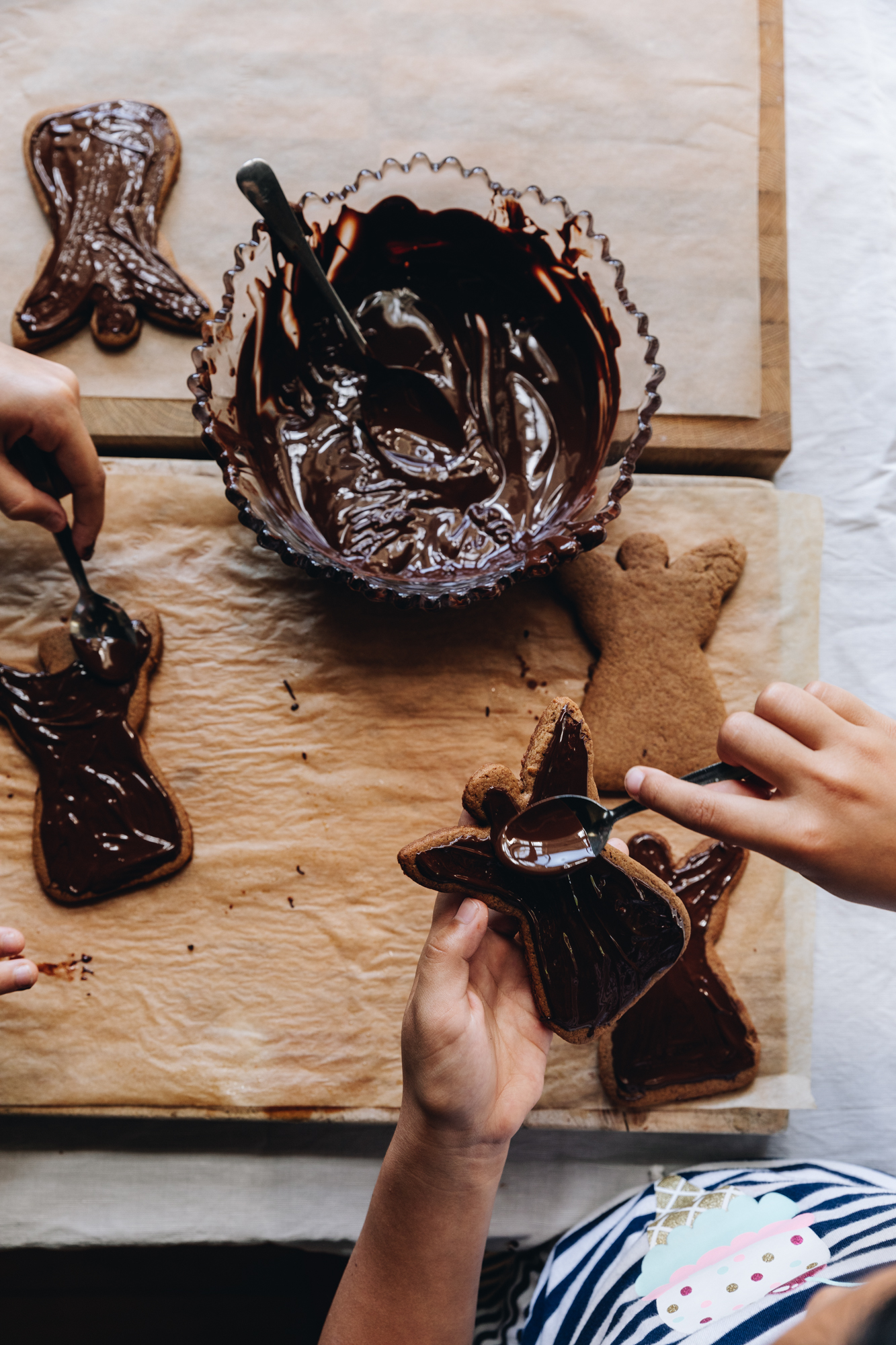 A flat lay shot shows spiced gingerbread cookies that are shaped as angels being smeared with dark chocolate by children's hands. A vintage bowl has dark chocolate in it and sits on top of two wooden boards lined with brown baking paper.