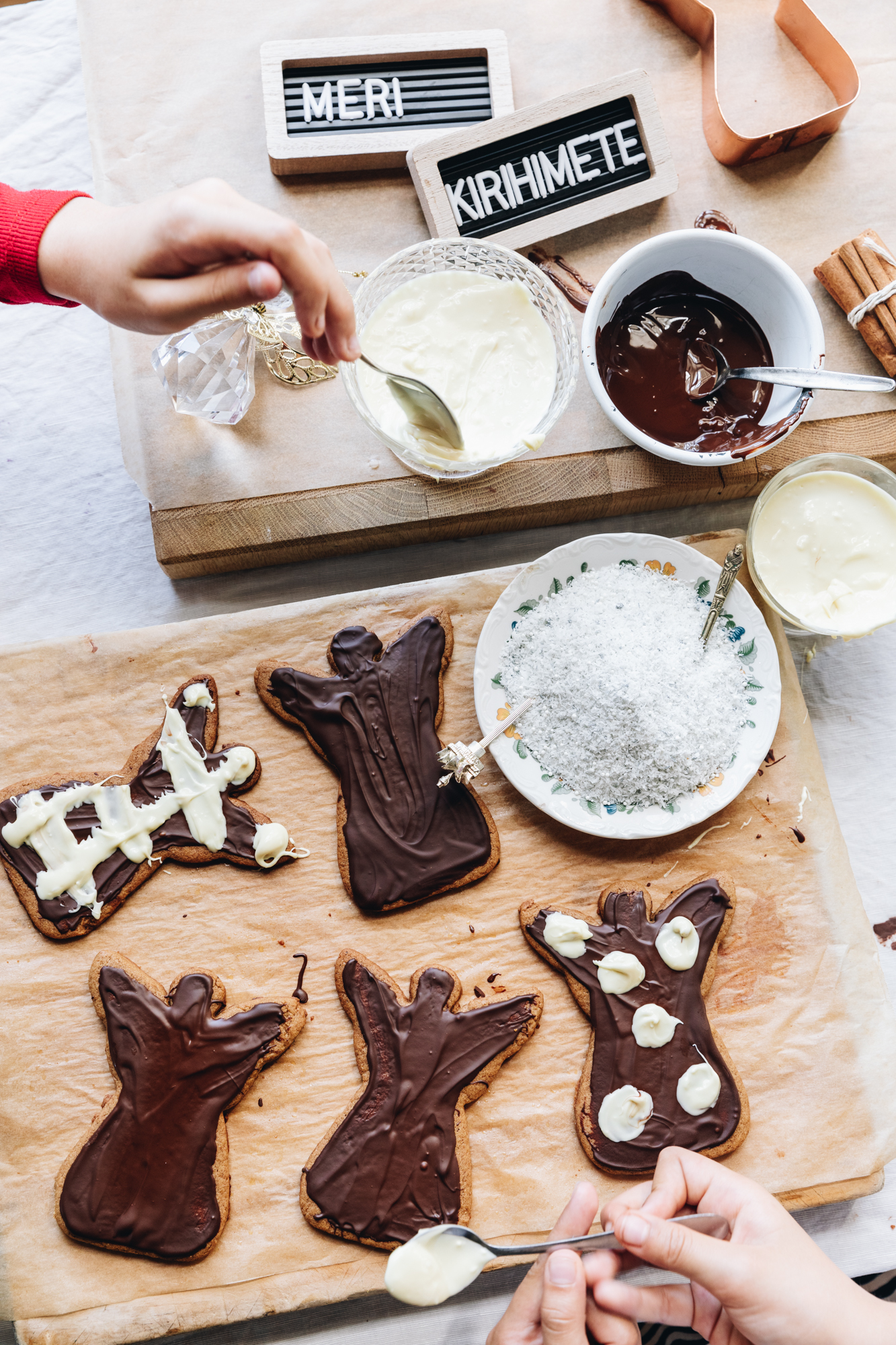 Two wooden boards are lined with brown baking paper. A flat lay shot shows spiced gingerbread cookies shaped as angels with dark chocolate on them. One Childs hand is getting melted white chocolate and the other is holding a teaspoon of white chocolate. Two of the gingerbread angels have white chocolate on them. A sign saying Meri Kirihimete is seen too. A plate of coconut is on the board and a small bowl of melted dark chocolate. 