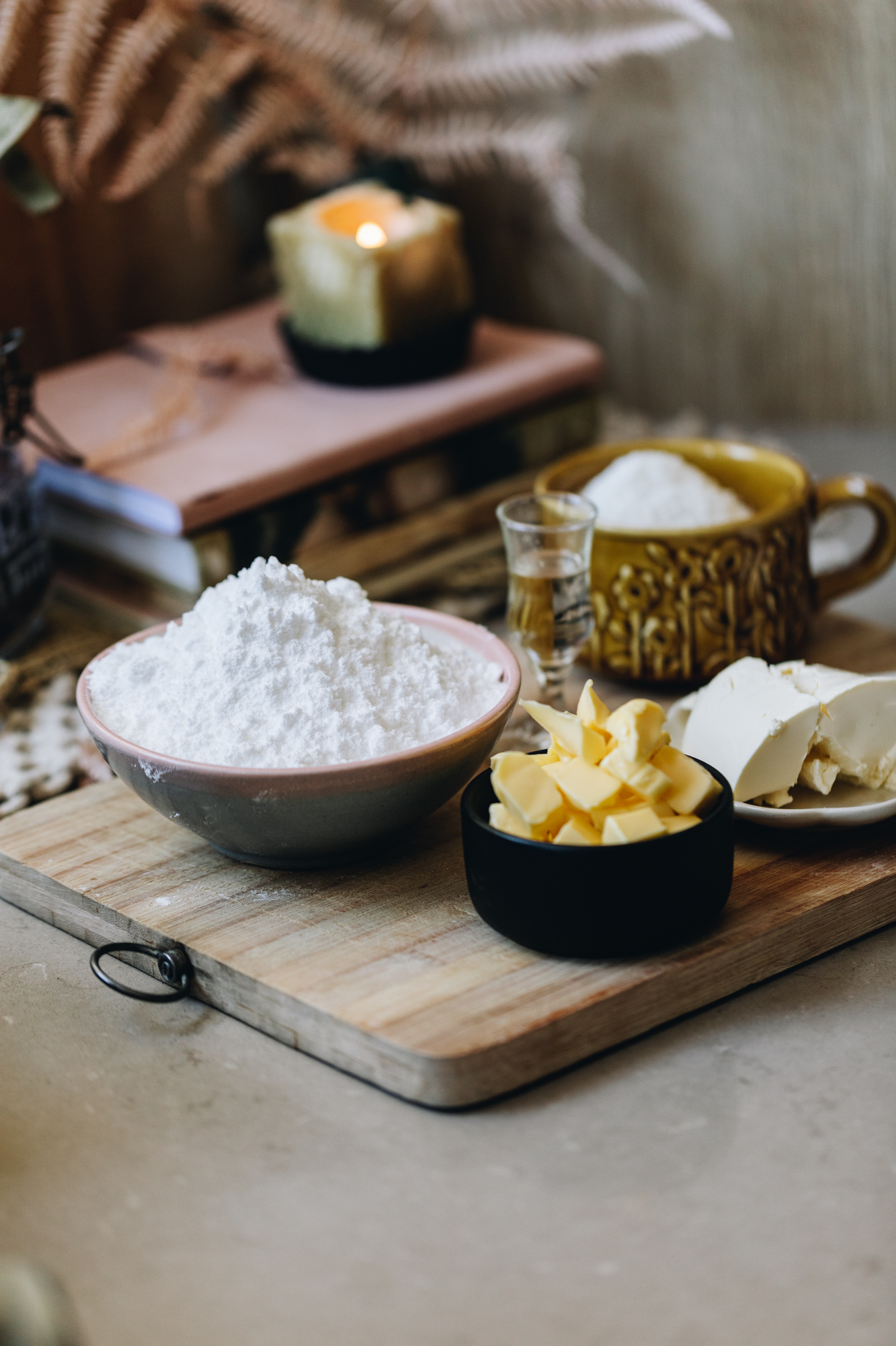 A wooden board sits on a brown stone bench. On the board is ingredients in vintage bowls. There is butter, cream cheese, icing sugar and peppermint essence in view. Behind the board is a pile of books with a small square candle burning. Behind that is pink dried flowers. 