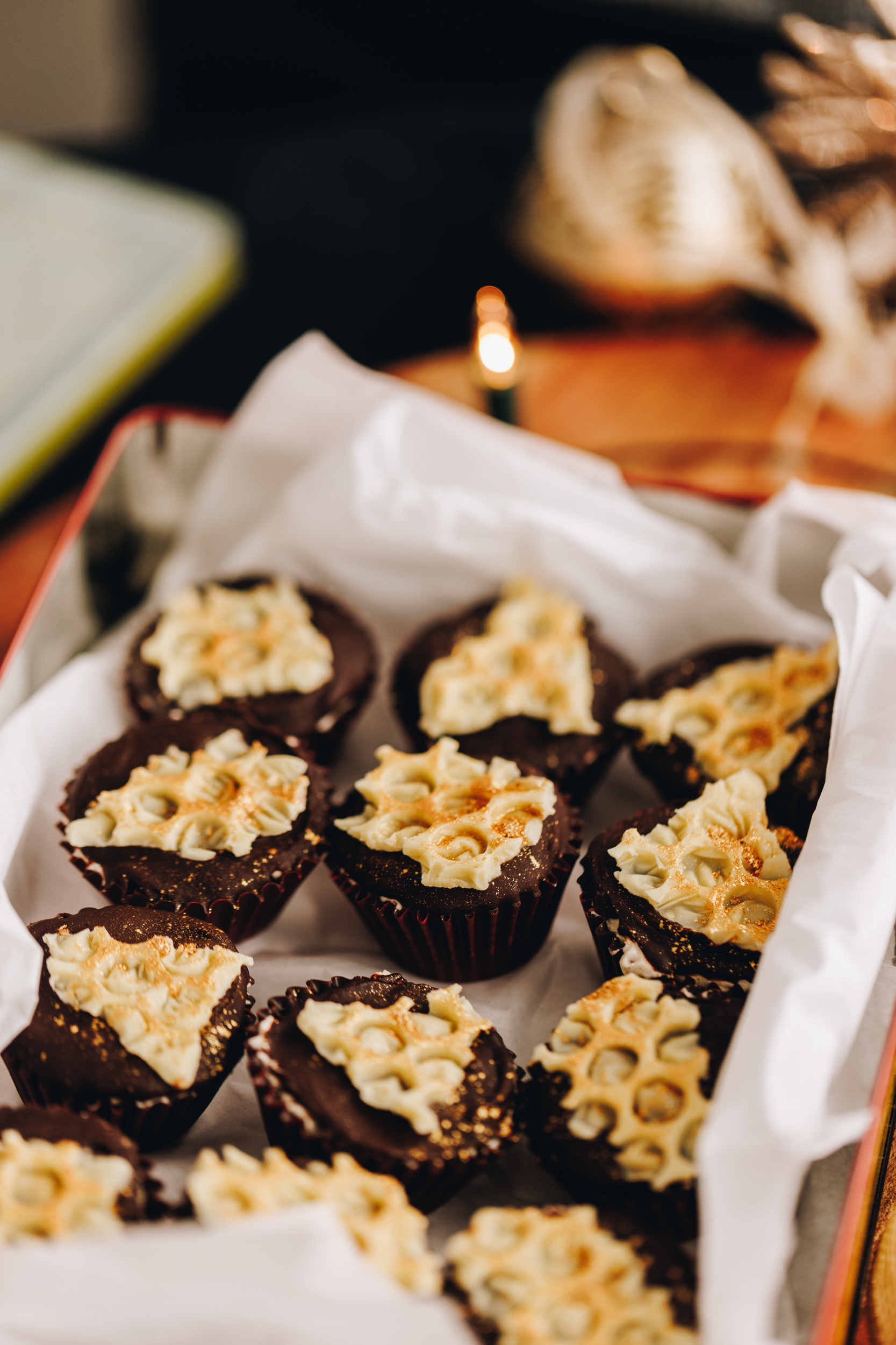 No-bake creamy peppermint cups are set and are in a cookie tin lined with white baking paper. A candle burns in the background.  The cups have a white chocolate bubble wrap decoration on each one dusted with edible gold dust. 