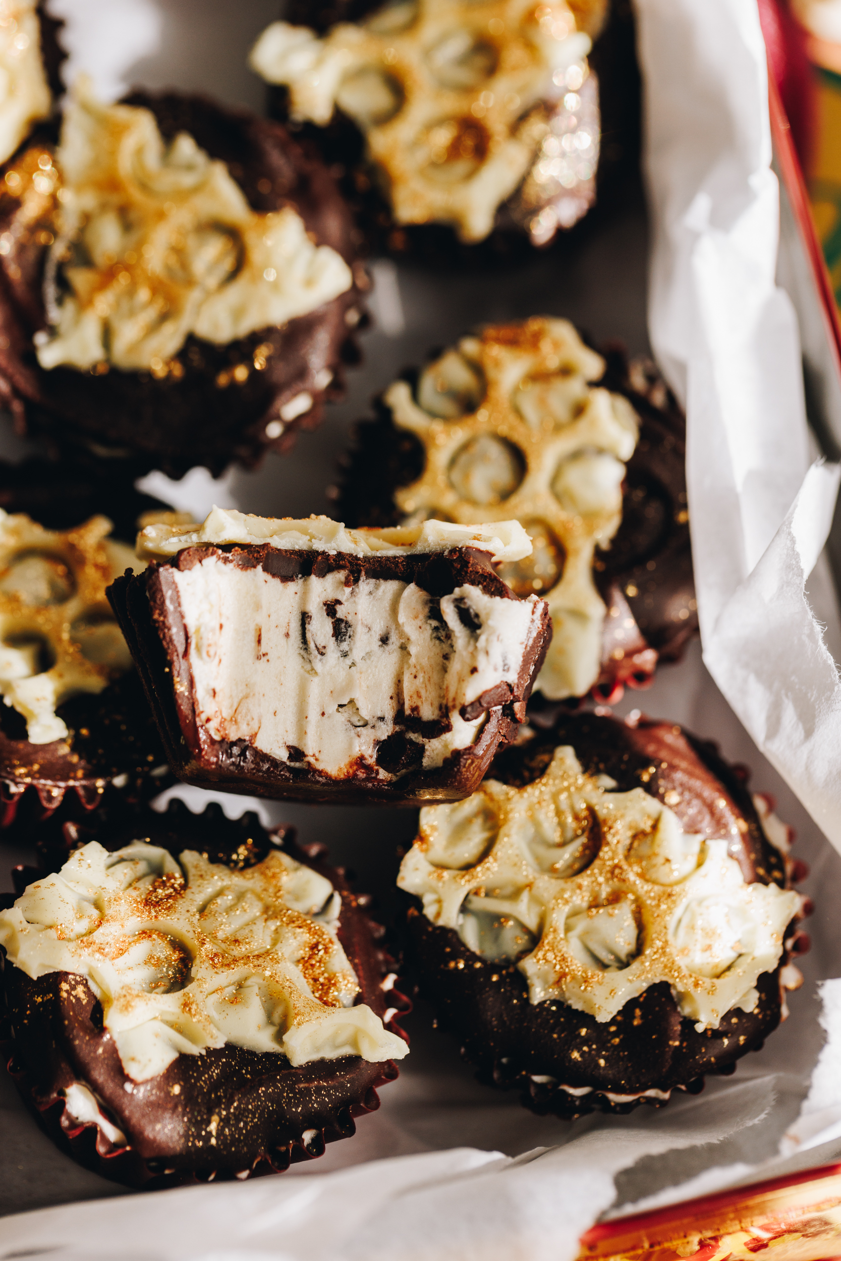 No-bake creamy peppermint cups are set and are in a cookie tin lined with white baking paper. The cups have a white chocolate bubble wrap decoration on each one dusted with edible gold dust. One of the cups have been bitten in to showing the creamy filling with chocolate pieces in it. 