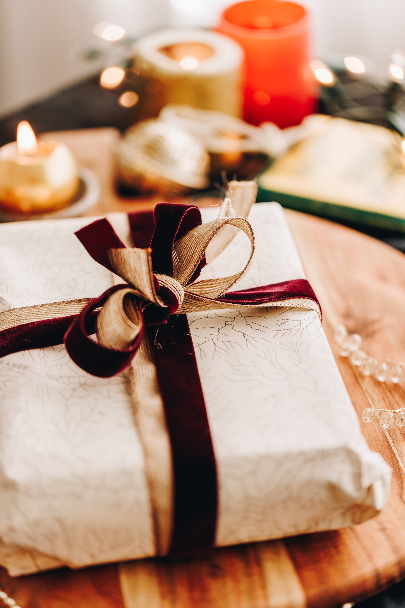 A rectangle gift is wrapped in a white paper with a map all over it. It is wrapped in a red velvet and beige natural fabric bow. Candle are burning in the background. 