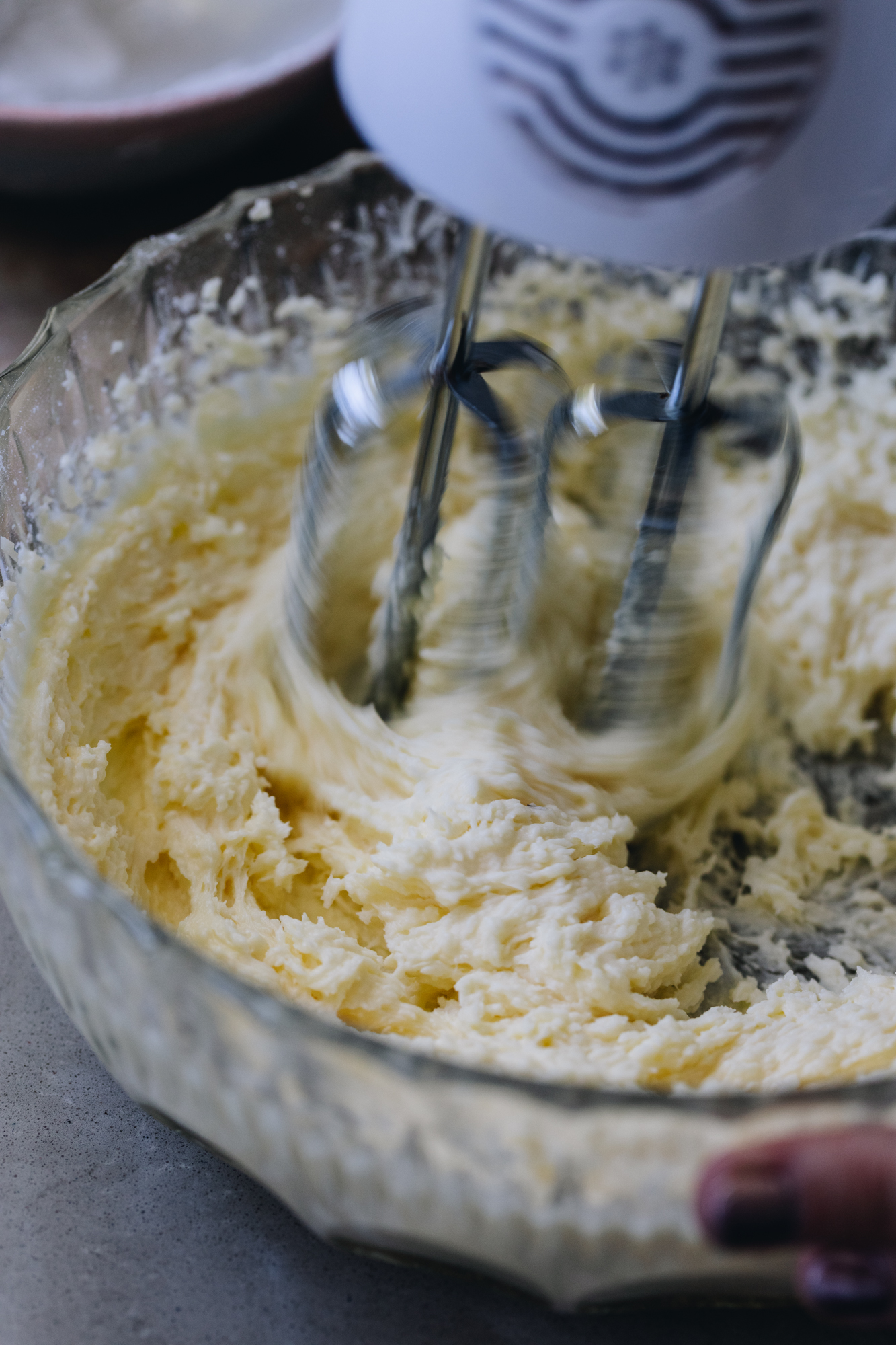 A vintage glass bowl sits on a brown bench. In the bowl is whipped cream cheese that is being whipped by a white hand mixer. 