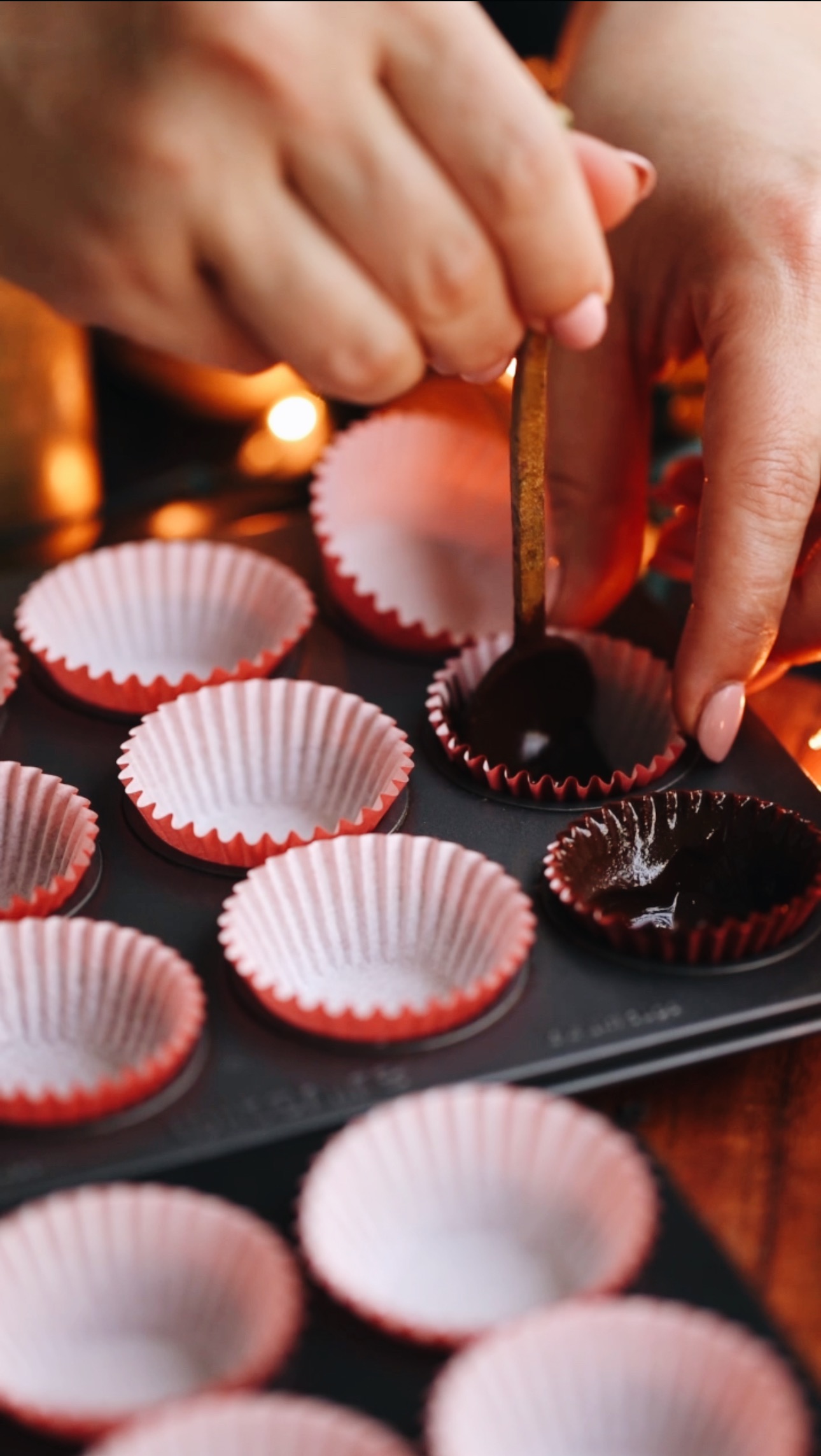 A mini muffin tin sits on a wooden board. It is lined with red cupcake cases and Naomi is spreading dark chocolate in to on elf the cases. Candles burn in the background.