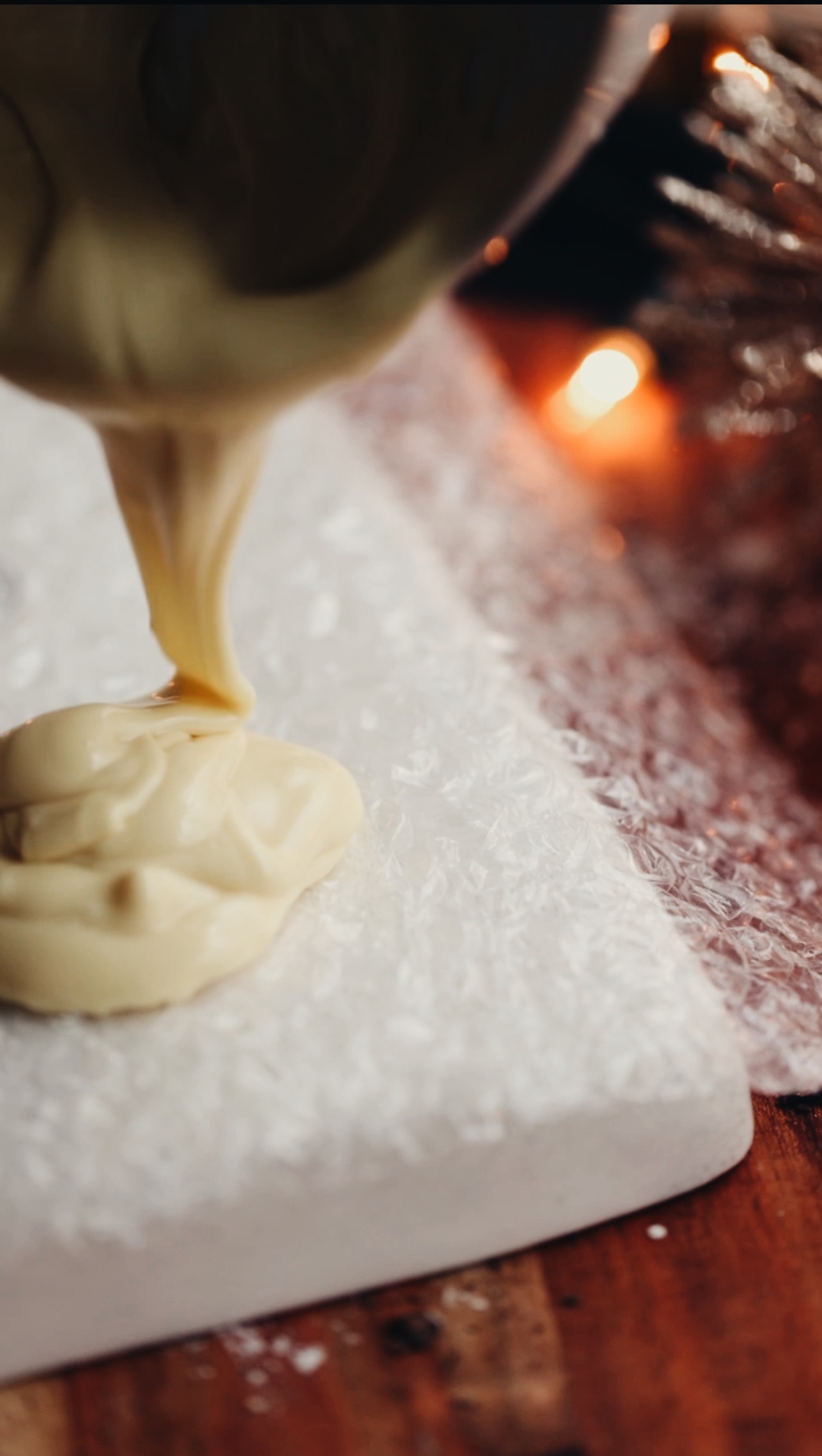Bubble wrap is on a white board that sits on a wooden table. Naomi is pouring melted white chocolate on to the bubble wrap from a bowl. Candles are burning in the background. 