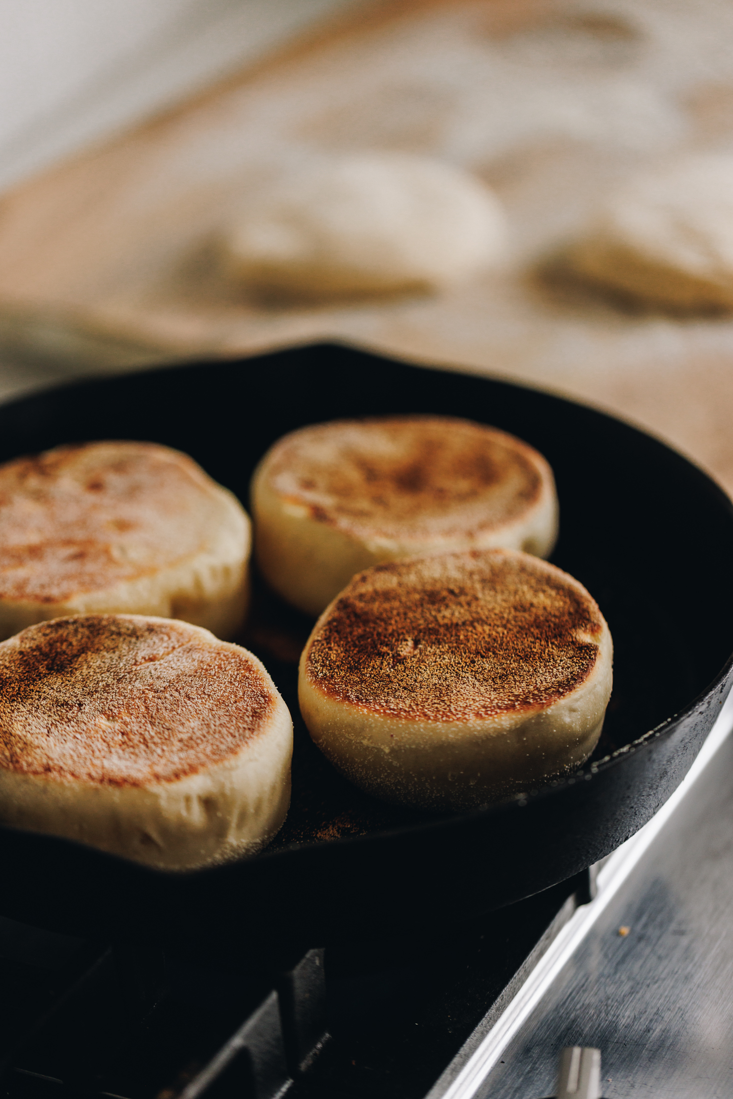 In a black cast iron pan is four homemade muffin splits cooking. The tops of a deep golden colour and in the background is a tray lined with brown baking paper and unbaked muffin split dough on it. 