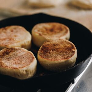 In a black cast iron pan is four homemade muffin splits cooking. The tops of a deep golden colour and in the background is a tray lined with brown baking paper and unbaked muffin split dough on it.