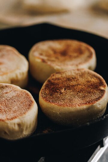 In a black cast iron pan is four homemade muffin splits cooking. The tops of a deep golden colour and in the background is a tray lined with brown baking paper and unbaked muffin split dough on it.