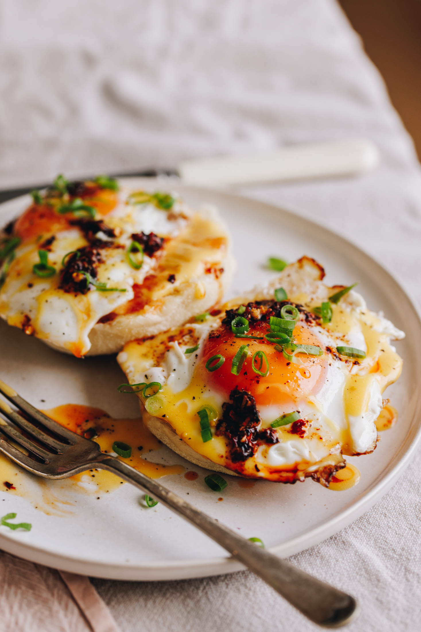 On a table is a white tablecloth with a white plate and a silver fork. On the plate is two fried eggs on muffin splits with homemade hollandaise sauce, chill oil and spring onions on top.