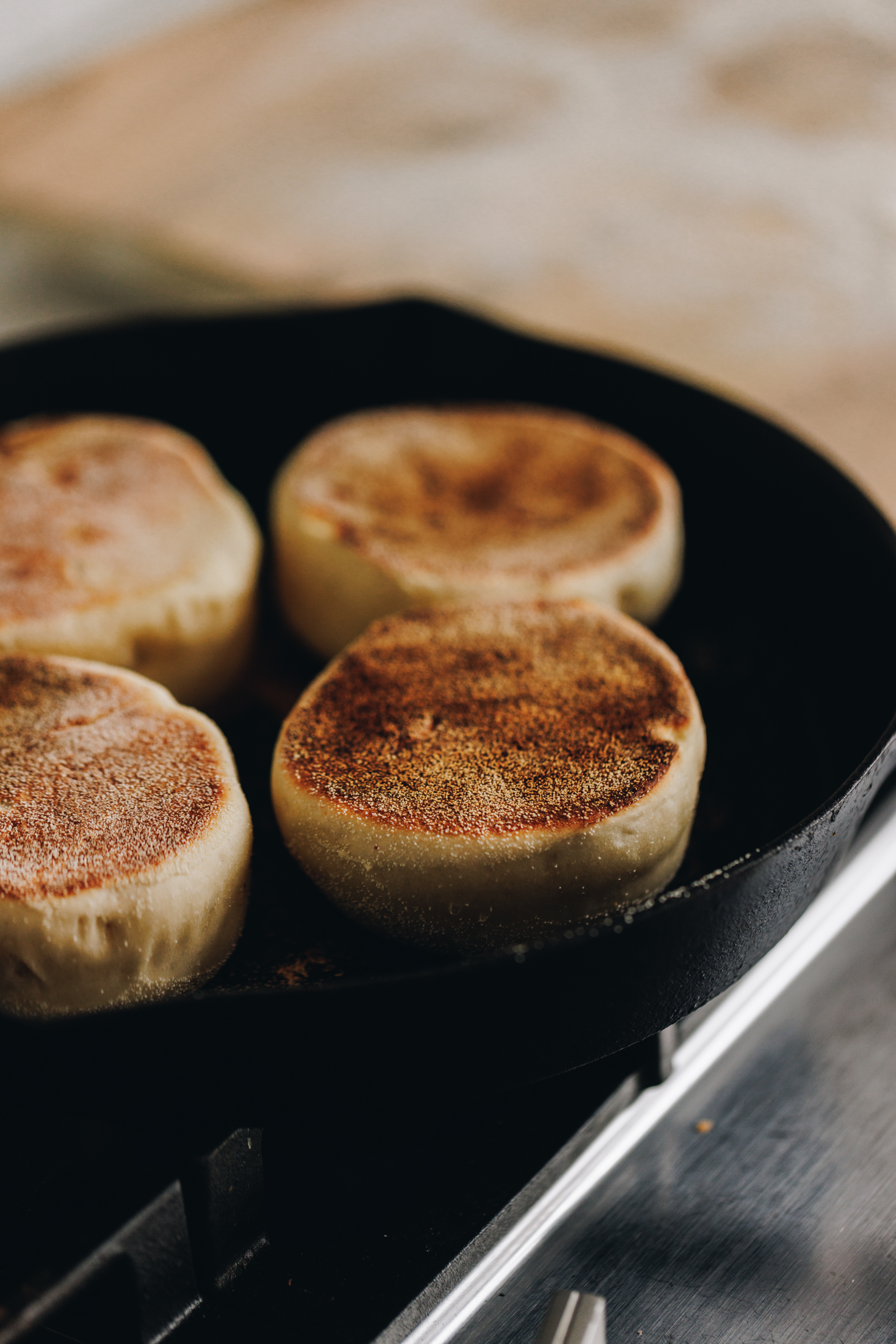 In a black cast iron pan is four homemade muffin splits cooking. The tops of a deep golden colour and in the background is a tray lined with brown baking paper.