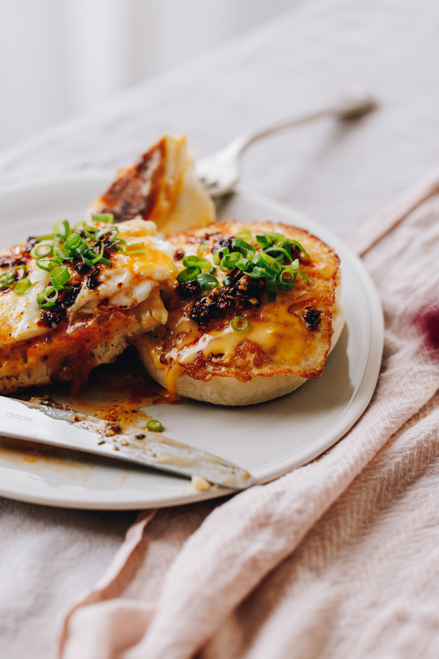 On a table is a white tablecloth with a white plate and a silver fork. A soft tablecloth is also under the plate. On the plate is two fried eggs on muffin splits with homemade hollandaise sauce, chill oil and spring onions on top. A knife is on the plate and a fork is behind it. 