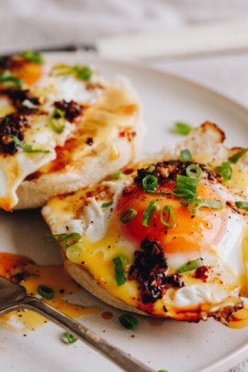 On a table is a white tablecloth with a white plate and a silver fork. On the plate is two fried eggs on muffin splits with homemade hollandaise sauce, chill oil and spring onions on top.