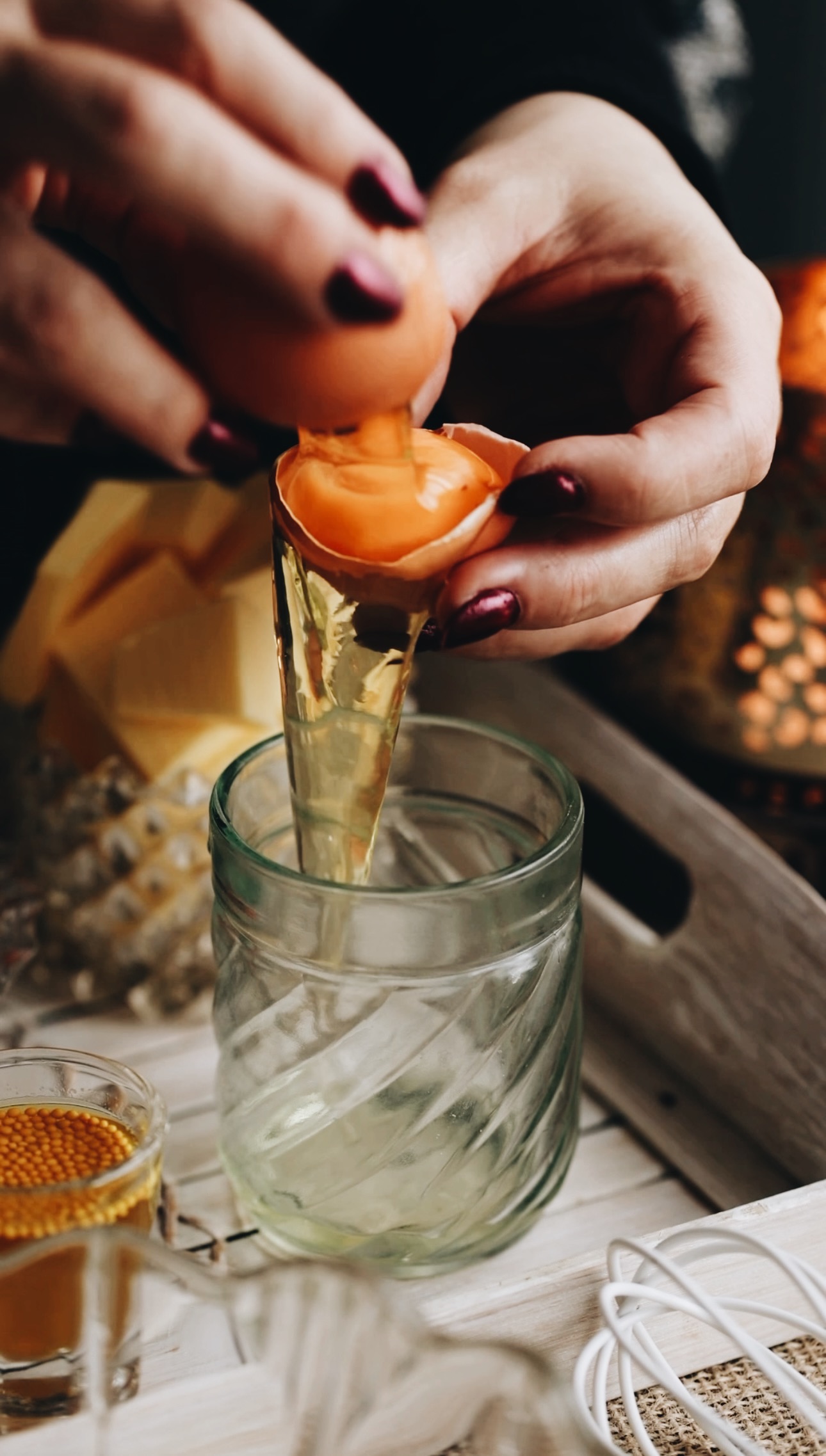 A vintage glass is on a table. In the glass egg whites are falling in to it as Naomi catches the egg yolk in the egg shells. Next to the glass is mustard seeds in a juice. A white tray is beside it too and a glass jar of chopped up butter is behind it. 