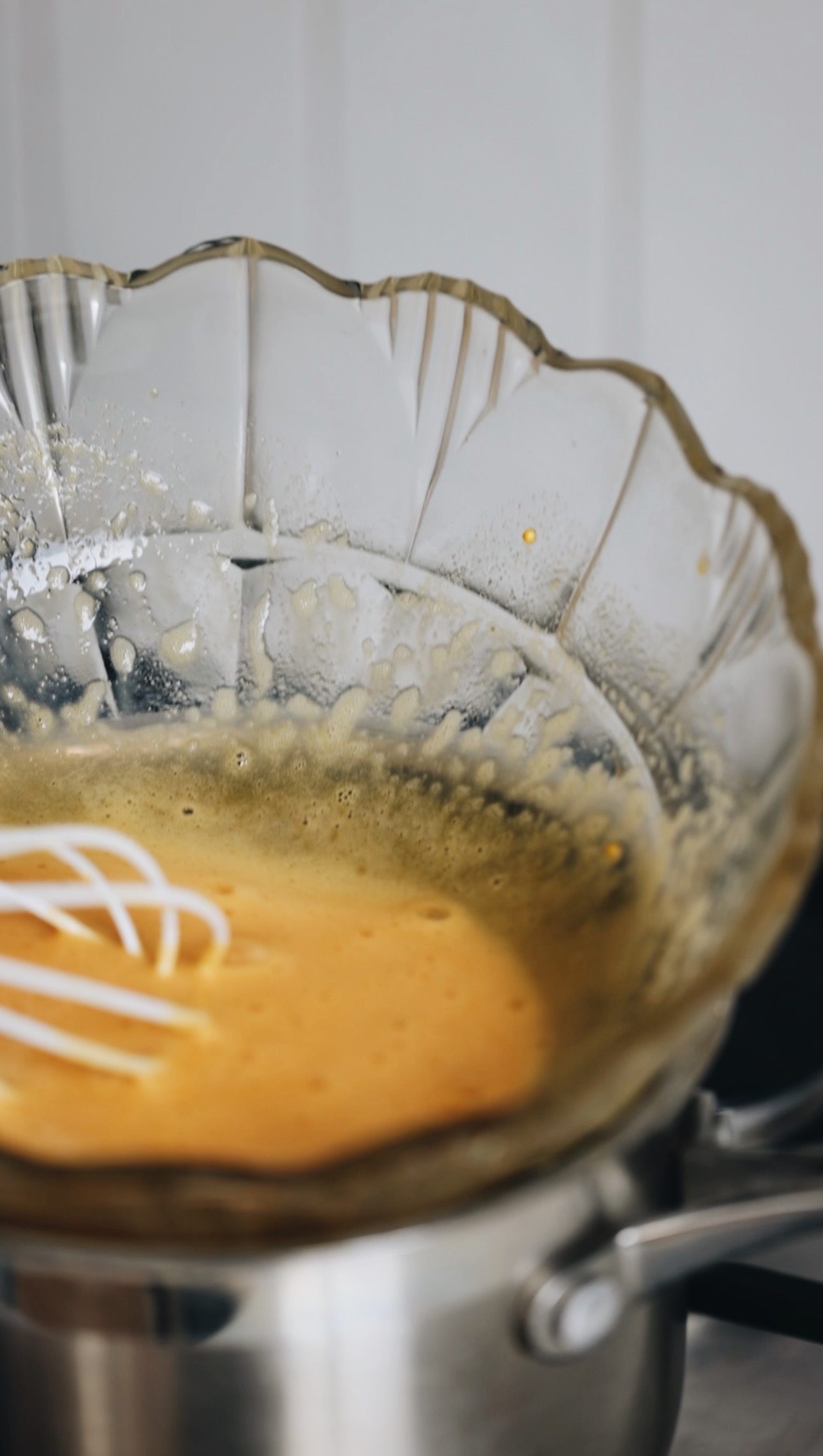 A silver pot sits on a stove with a vintage glass bowl on top of it. In the bowl is a pale egg yolk mixture that is being stirred with a white whisk.
