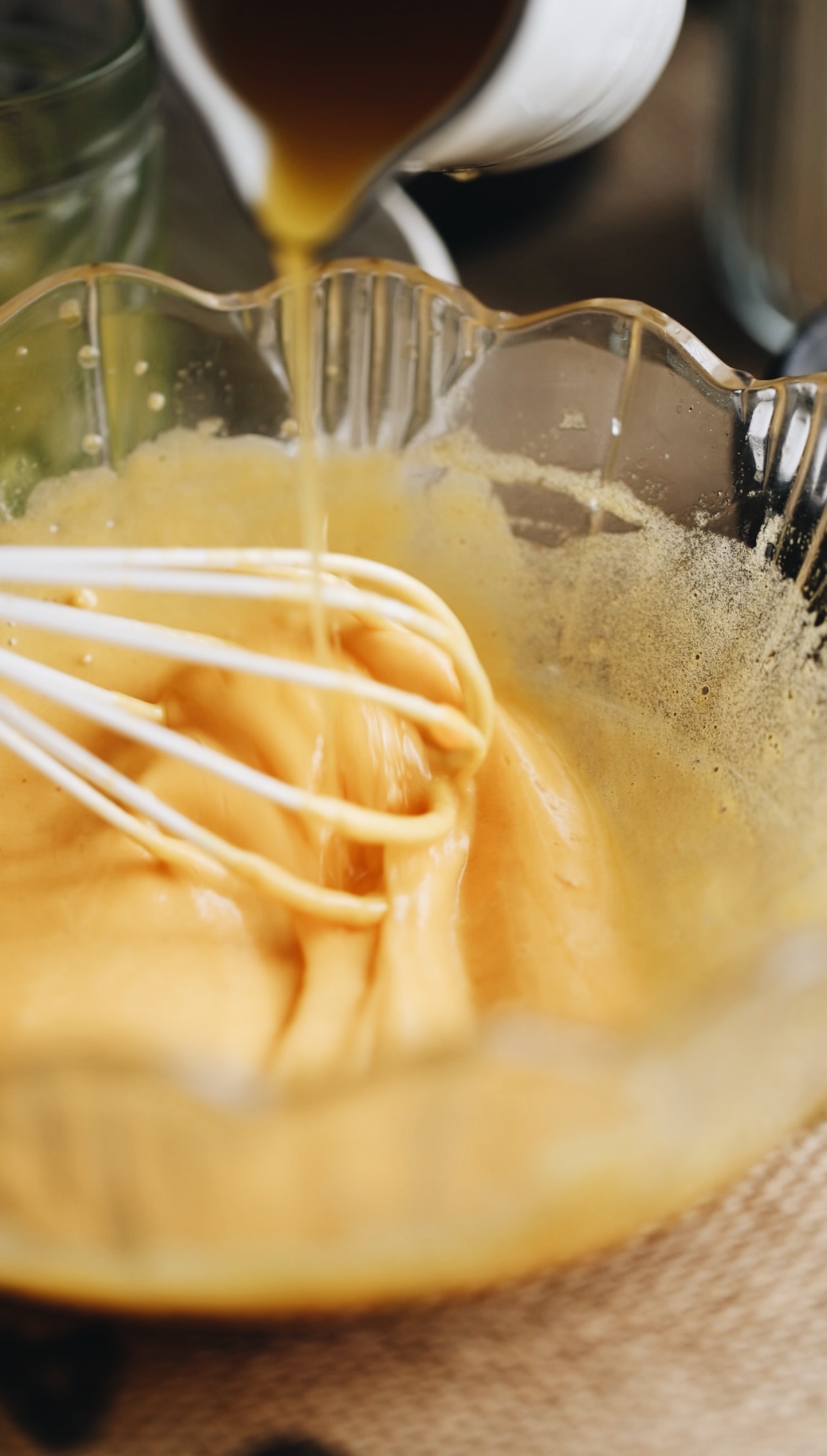 A glass vintage bowl has a thick egg yolk mixture in it and is being whisked with a white whisk. At the same time a white jug is pouring melted butter in to it. 