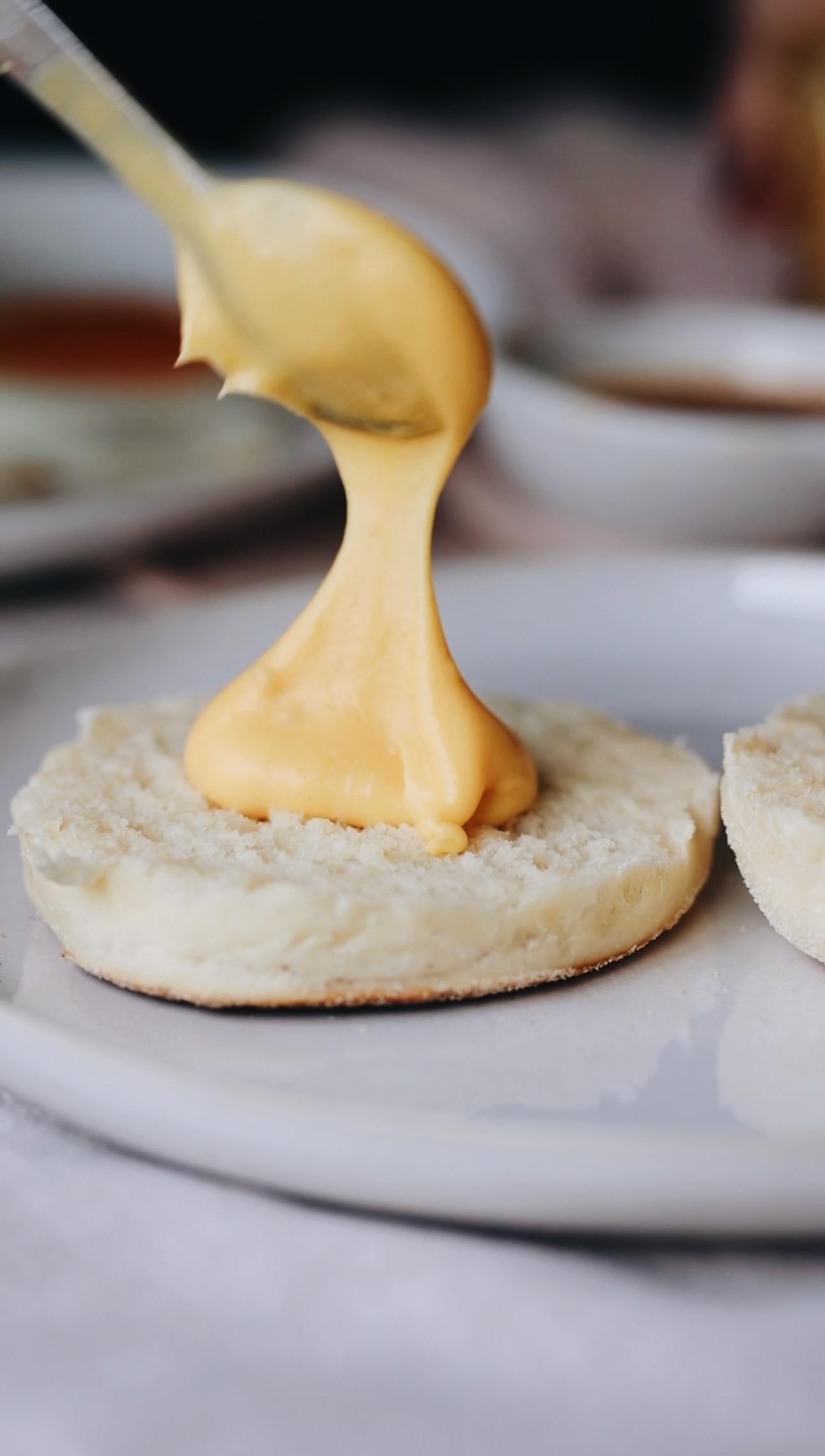A white plate sits on a grey tablecloth. On the plate is a piece of muffin split that has a large dollop of homemade hollandaise sauce being added to it with a teaspoon. 