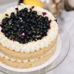 A whole pistachio and blueberry sponge sits on a glass cake stand. In the centre of the sponge is piped cream. Piped cream decorates the top of the cake and in the centre is a blueberry compote, fresh blueberries and flower petals.