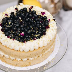A whole pistachio and blueberry sponge sits on a glass cake stand. In the centre of the sponge is piped cream. Piped cream decorates the top of the cake and in the centre is a blueberry compote, fresh blueberries and flower petals.