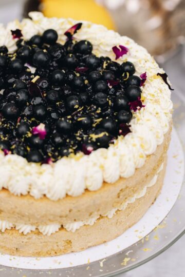 A whole pistachio and blueberry sponge sits on a glass cake stand. In the centre of the sponge is piped cream. Piped cream decorates the top of the cake and in the centre is a blueberry compote, fresh blueberries and flower petals.