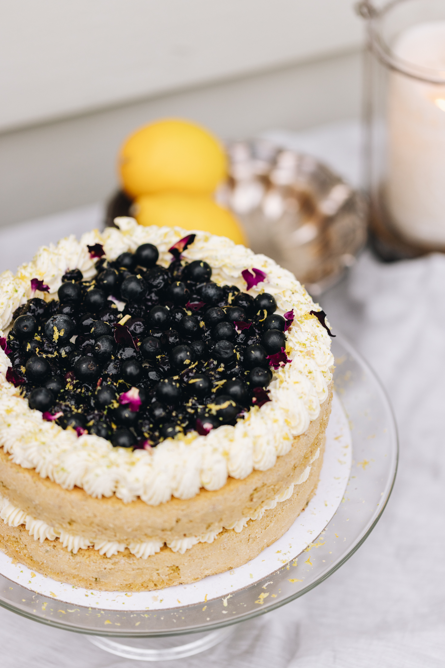 A whole pistachio and blueberry sponge sits on a glass cake stand. In the centre of the sponge is piped cream. Piped cream decorates the top of the cake and in the centre is a blueberry compote, fresh blueberries and flower petals. The cake stand sits on a grey table cloth outside with lemons and a candle in the background. 