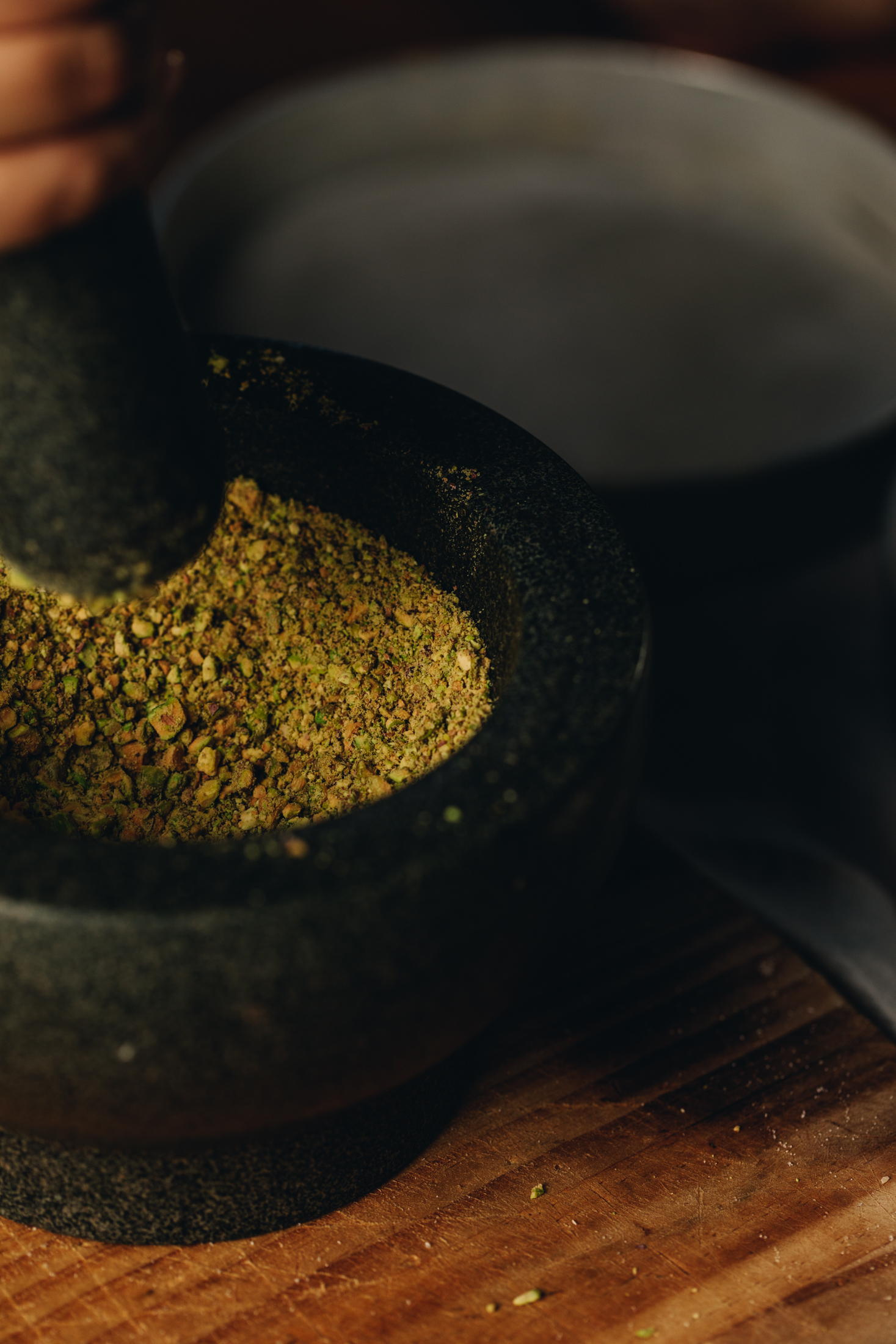 A dark mortar and pestle sits on a wooden table. In the mortar and pestle is crushed pistachios. A cake tin is in the background. 