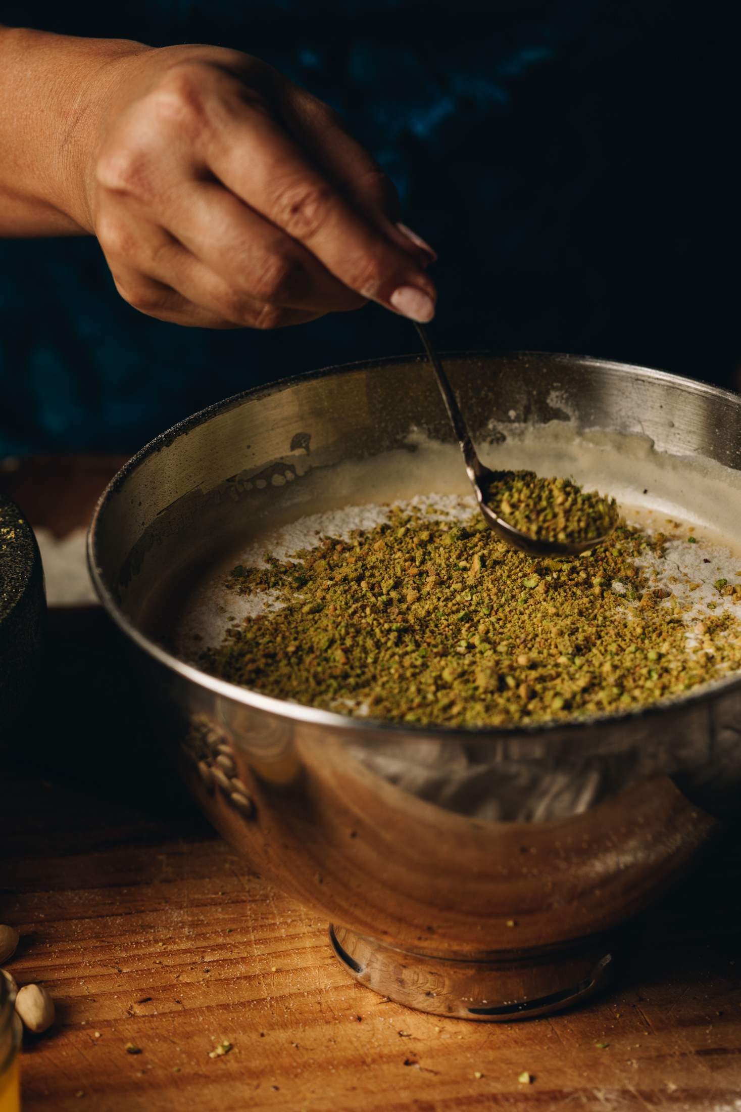 A silver mixing bowl sits on a wooden table. In the bowl is a mixture of whipped eggs that looks fluffy. On top of the mixture is sifted flour and Naomi is sprinkled over a pistachio crumb on top of that. 