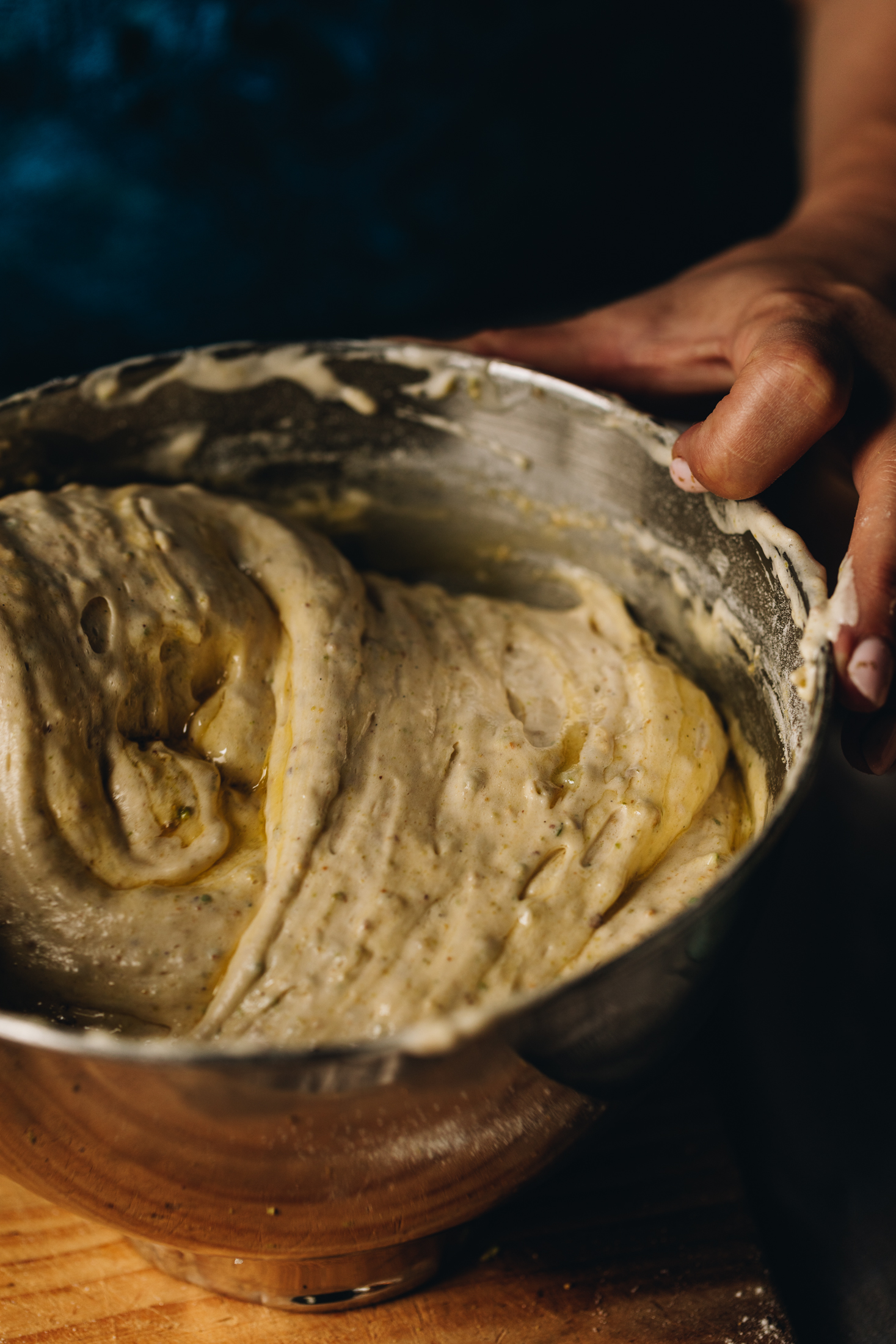 A silver mixing bowl sits on a wooden table. In the bowl is a mixture of whipped eggs that looks fluffy. The mixture is being folded and steaks of melted butter are seen. 