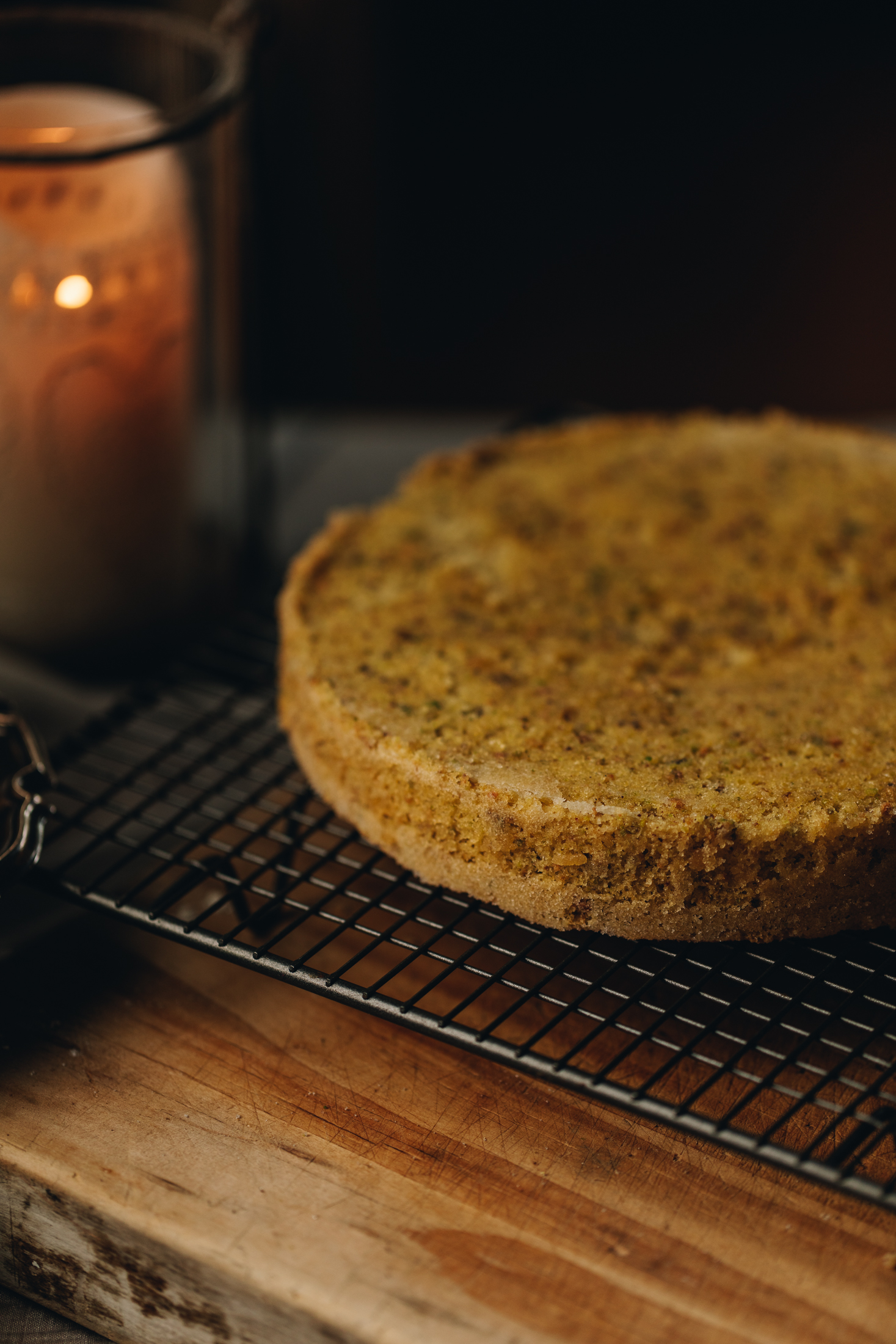 A baked pistachio and blueberry sponge is on a cooling rack that sits on a wooden table. A candle is burning next to it. 