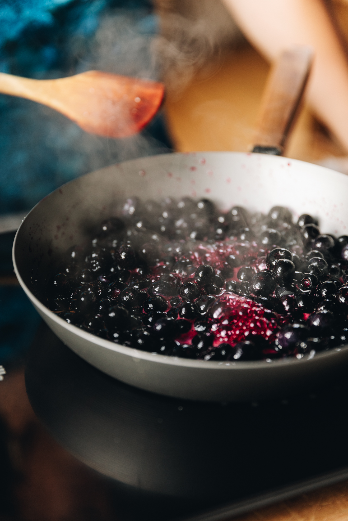 A silver frying pan with a wooden handle is on top of a black portable cook top. In the pan is blueberries that are cooking with steam coming off them. Naomi is holding a wooden spoon in the background. 