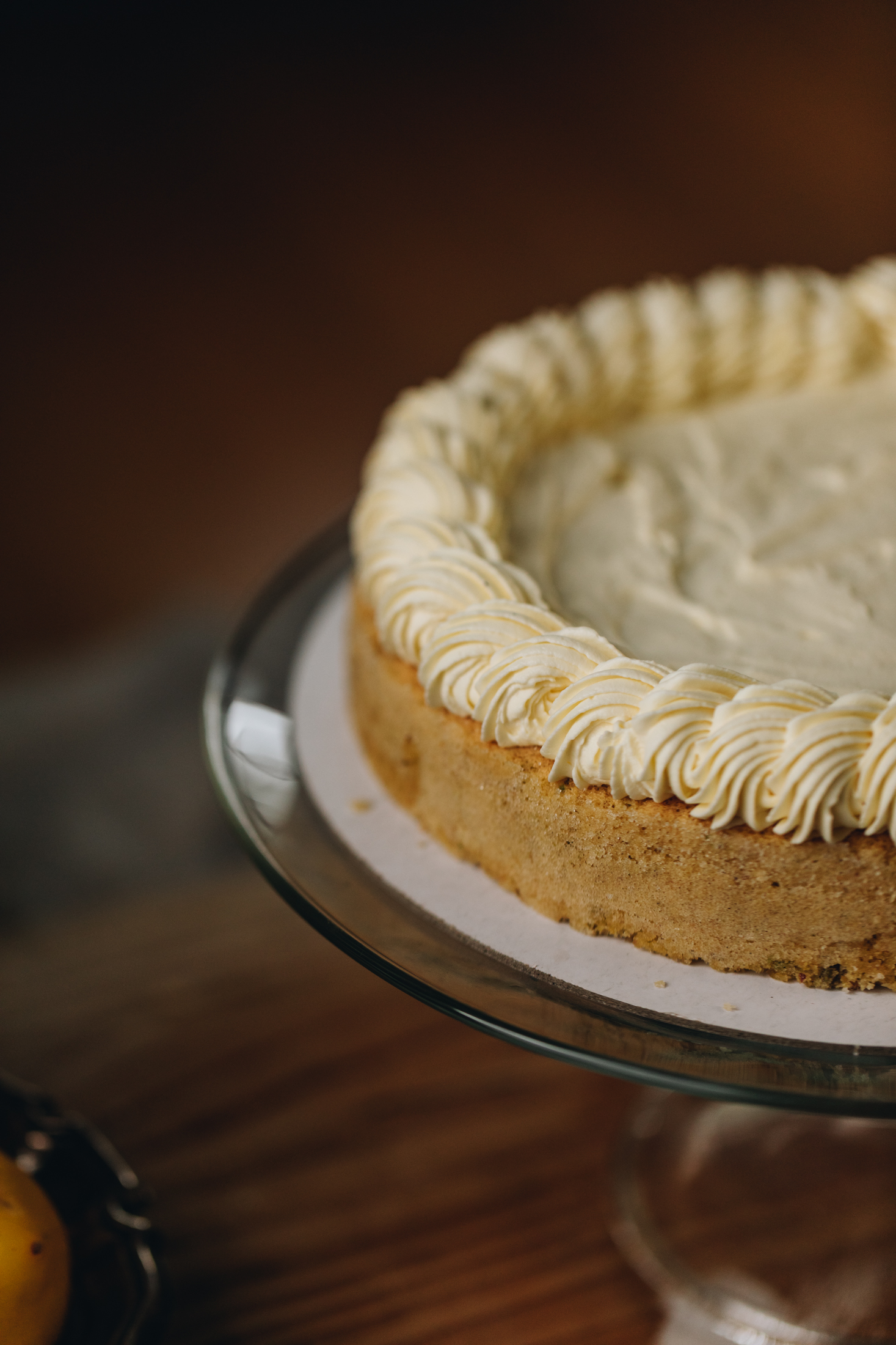 A sponge cake sits on a glass cake stand with cream spread on the top and piped cream around the edge. It is on a wooden table. 