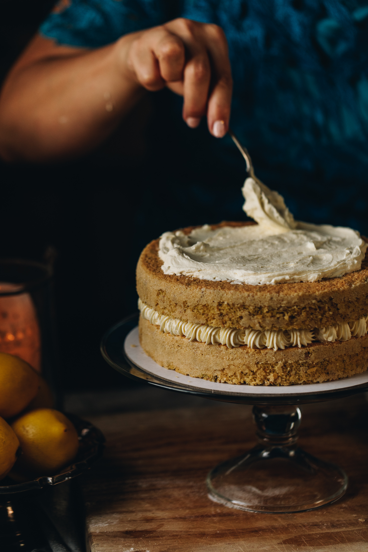 A sponge cake sits on a glass cake stand with cream spread on the top and piped cream around the edge. It has the lid on top and Naomi is spreading cream on top of it. It is on a wooden table.
