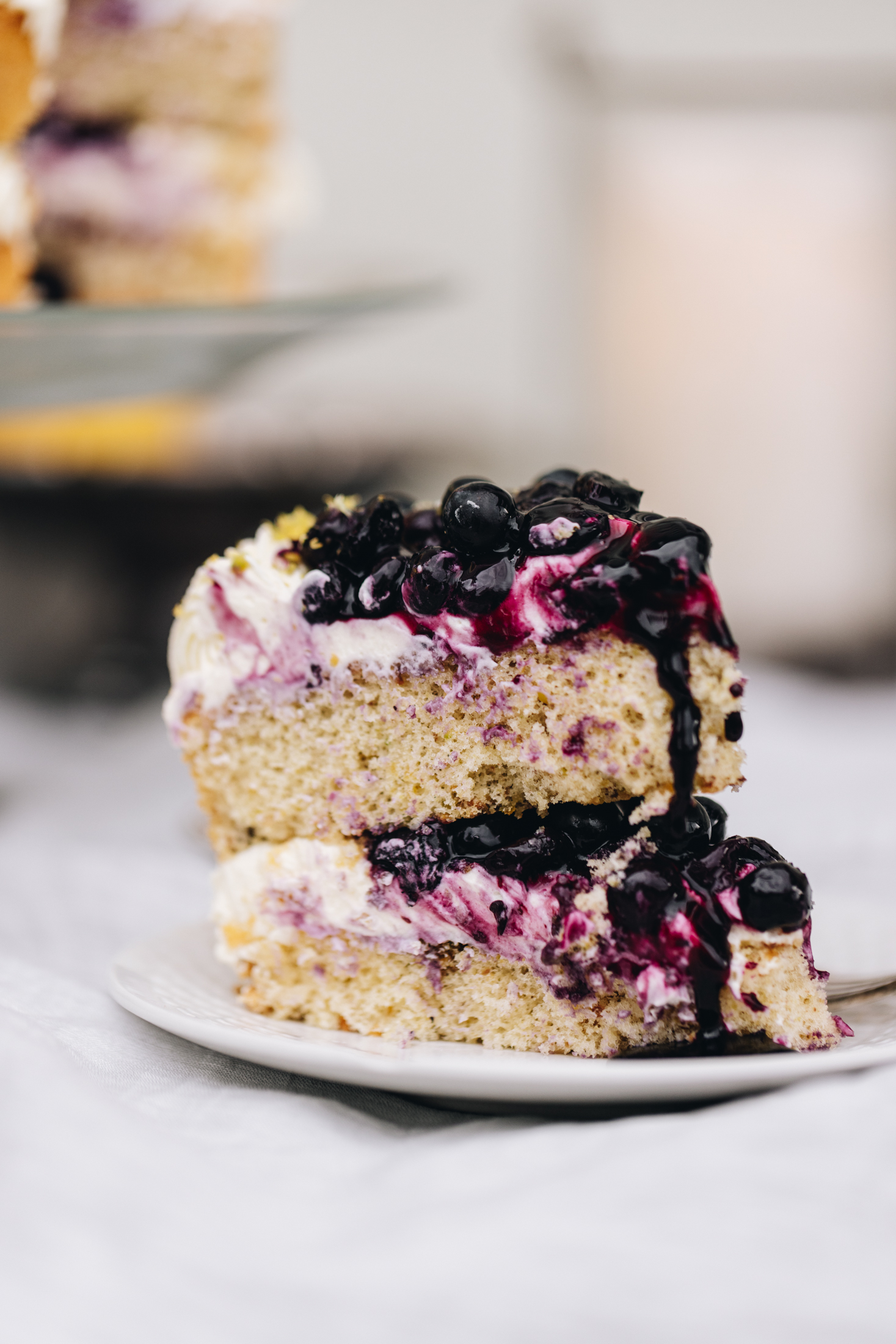 A slice of pistachio and blueberry sponge sits on a white plate. Blueberries and blueberry juice oozes out of the cake with fluffy cream.  