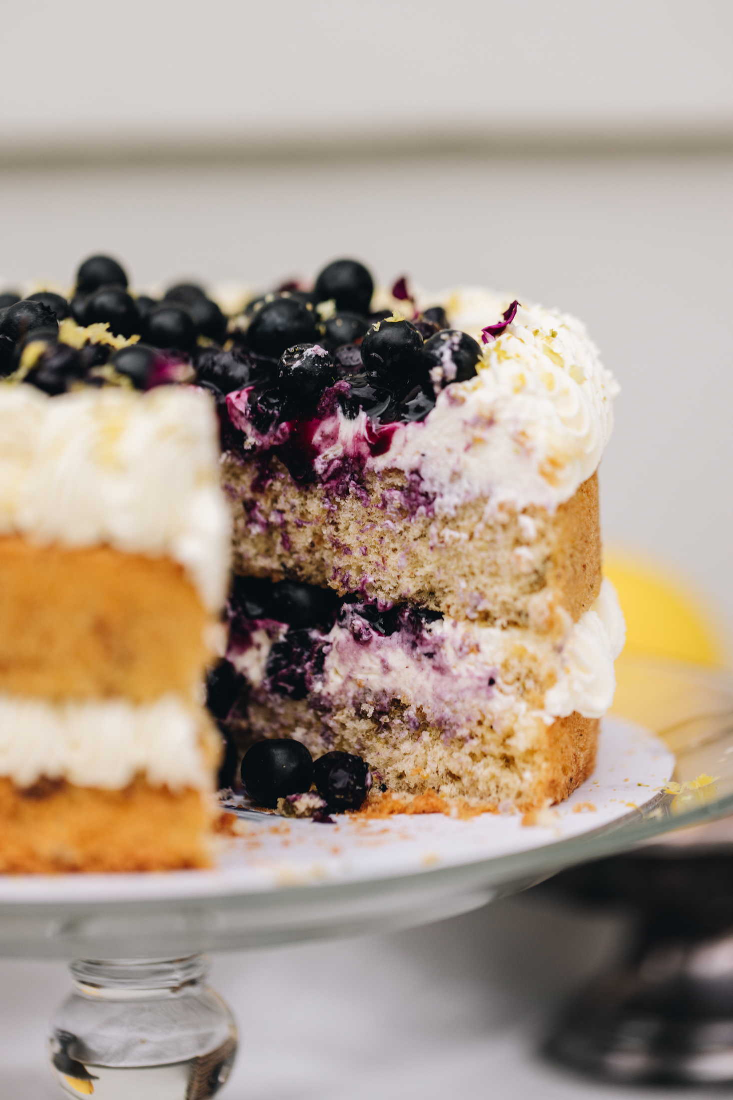A whole pistachio and blueberry sponge sits on a glass cake stand. In the centre of the sponge is piped cream. Piped cream decorates the top of the cake and in the centre is a blueberry compote, fresh blueberries and flower petals. The cake stand sits on a grey table cloth outside with lemons in the background. A slice has been cut out from it showing the cream and blueberry filling. 