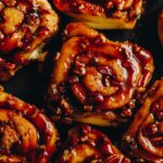 On a black tray sits sticky overnight maple cinnamon scrolls that have been pulled apart. They have sticky caramel on top of them and are studded with chopped pecan nuts.