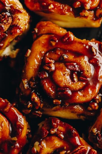 On a black tray sits sticky overnight maple cinnamon scrolls that have been pulled apart. They have sticky caramel on top of them and are studded with chopped pecan nuts.