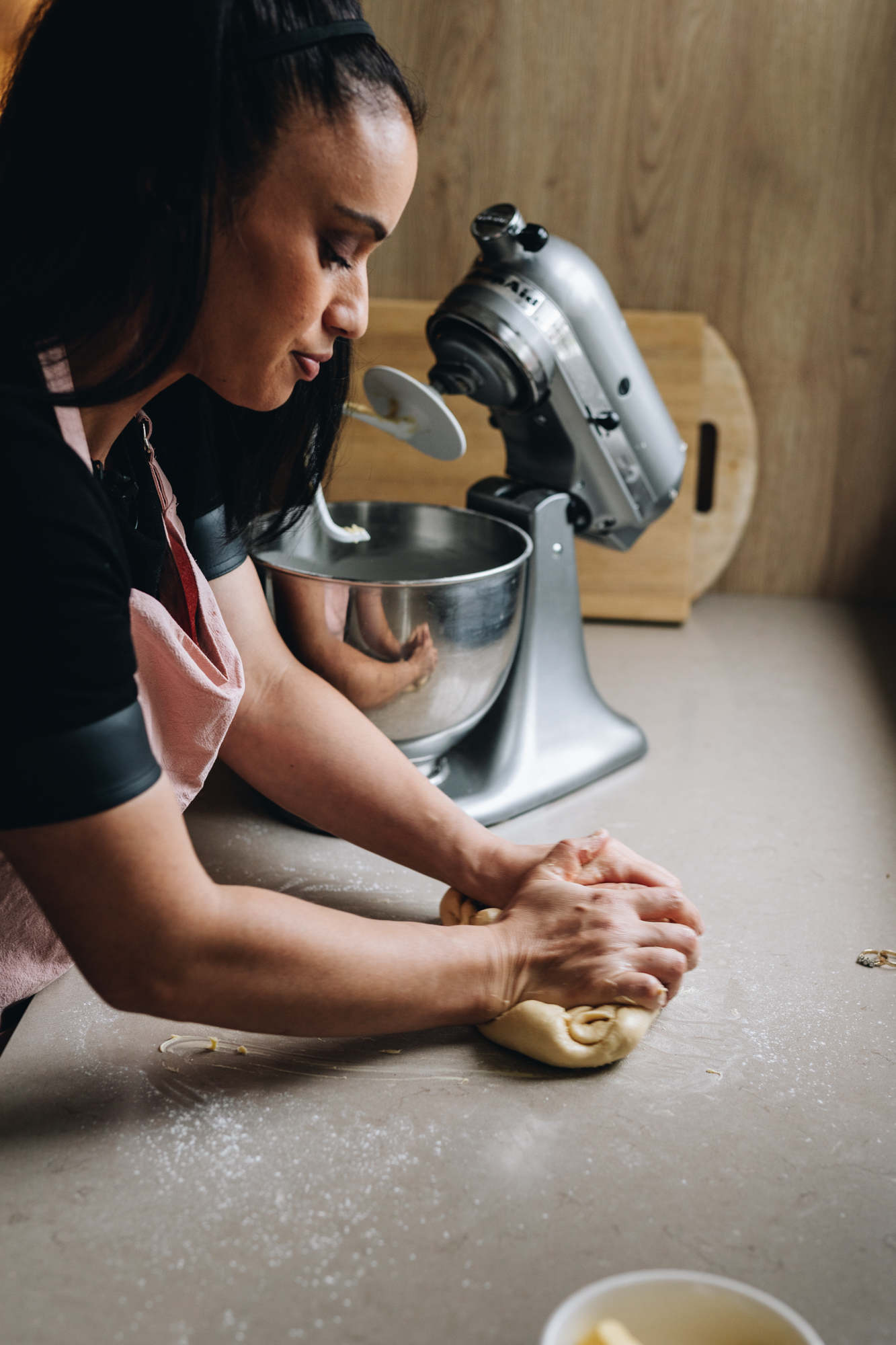 Naomi Toilalo is standing next to a brown stone bench. On the bench is a dough and she is kneading it with her hands. Behind her is a silver kitchen aid that has a dough hook attached. It is lifted up and the bowl is in the mixer too. There is a wooden board behind the mixer. 
