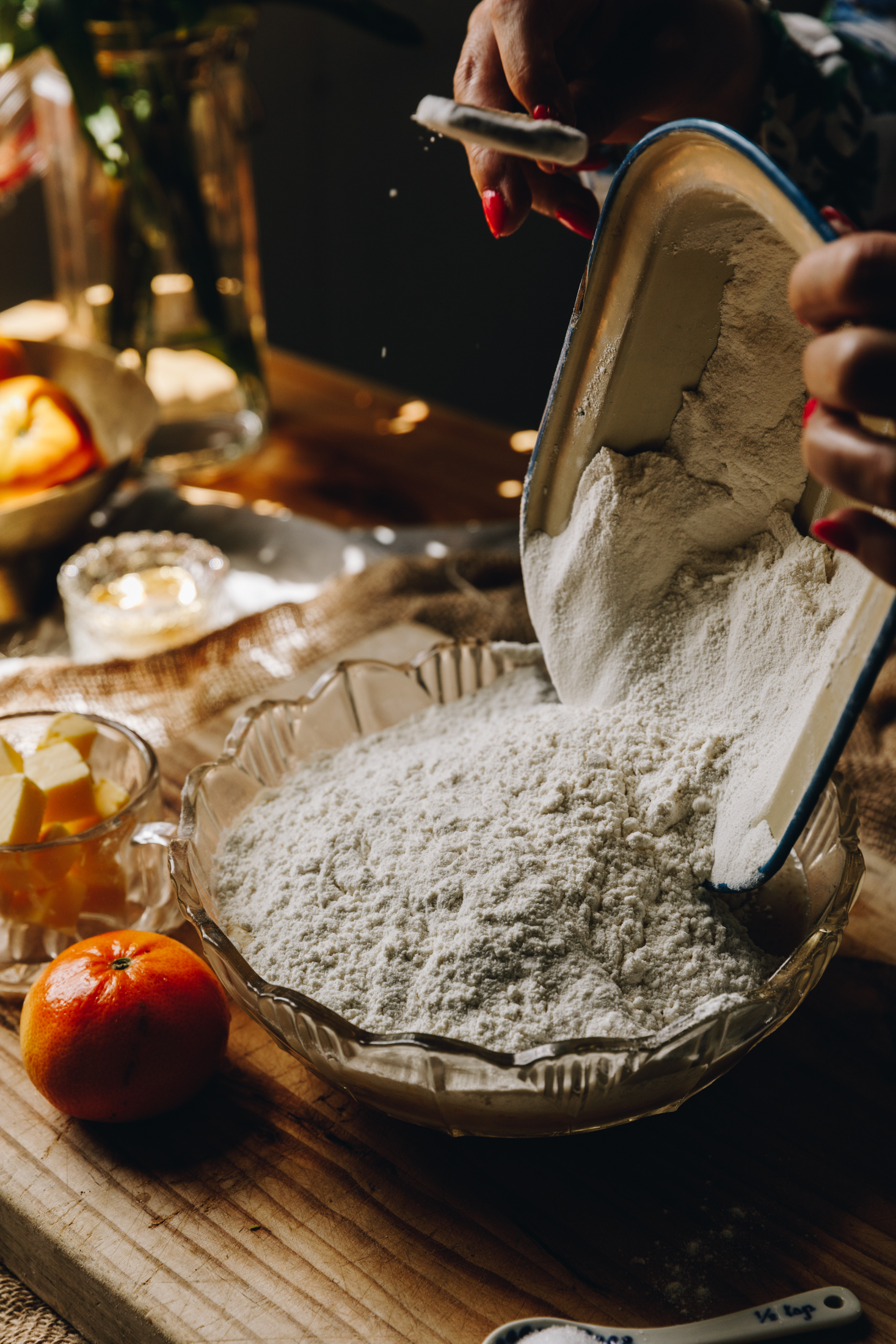 A vintage bowl sits on a wooden board. In the bowl is a large amount of flour and Naomi is tipping more flour in to it with a metal vintage dish. Next to the bowl is a mandarin,  a small glass of cubed butter and a candle. Behind is a glass vase with stems of flowers in it. 