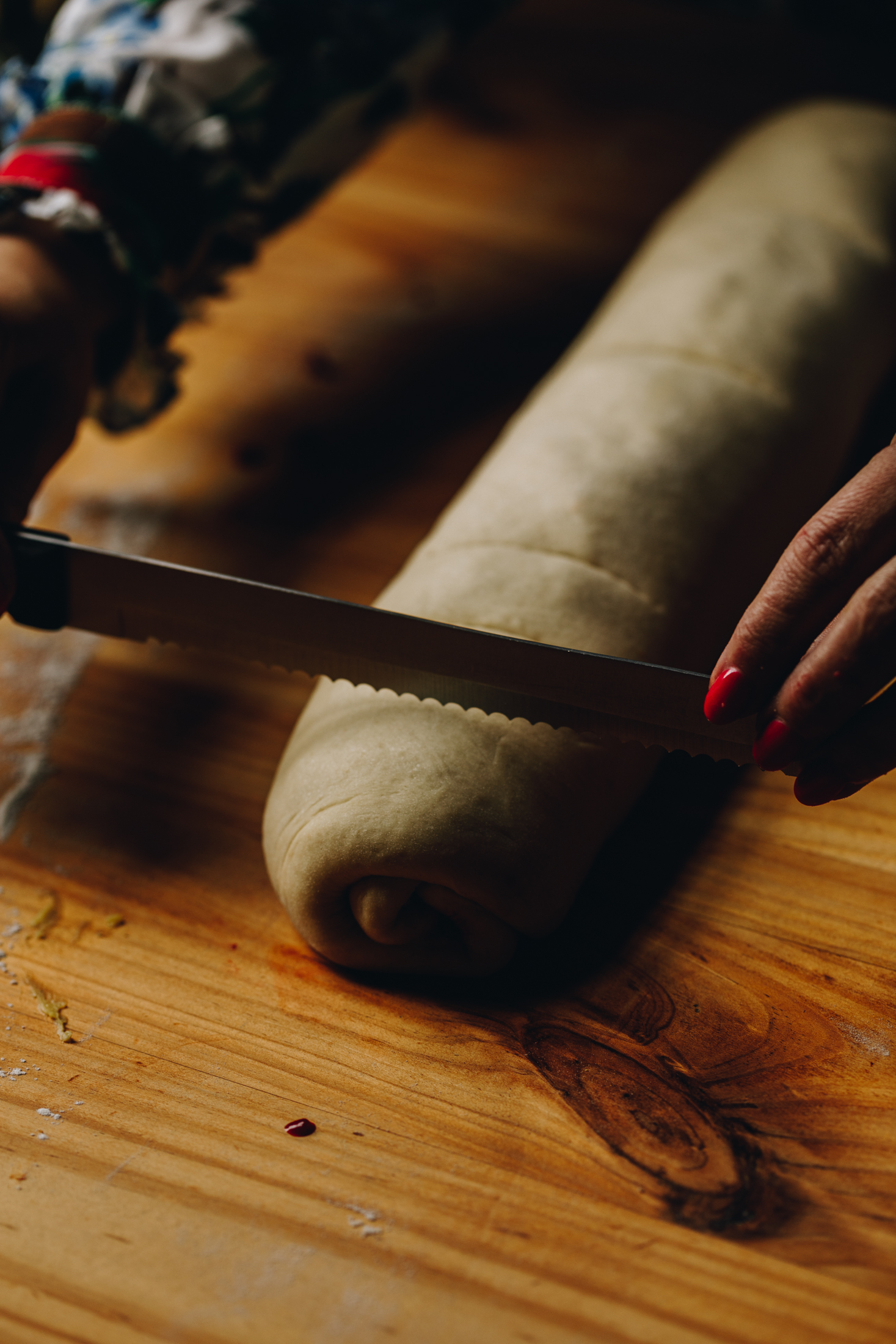 On a wooden table is a rolled up scroll dough. Naomi is marking out the sizes of the dough with a sharp knife.